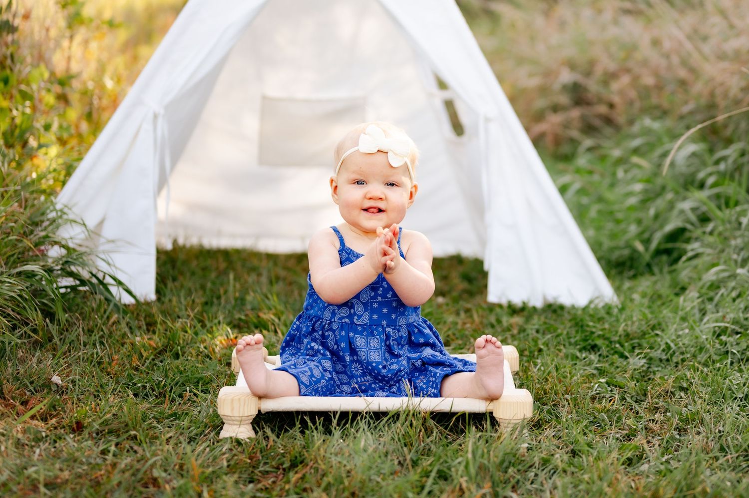 A happy toddler in a blue dress sits on a white platform near a teepee.