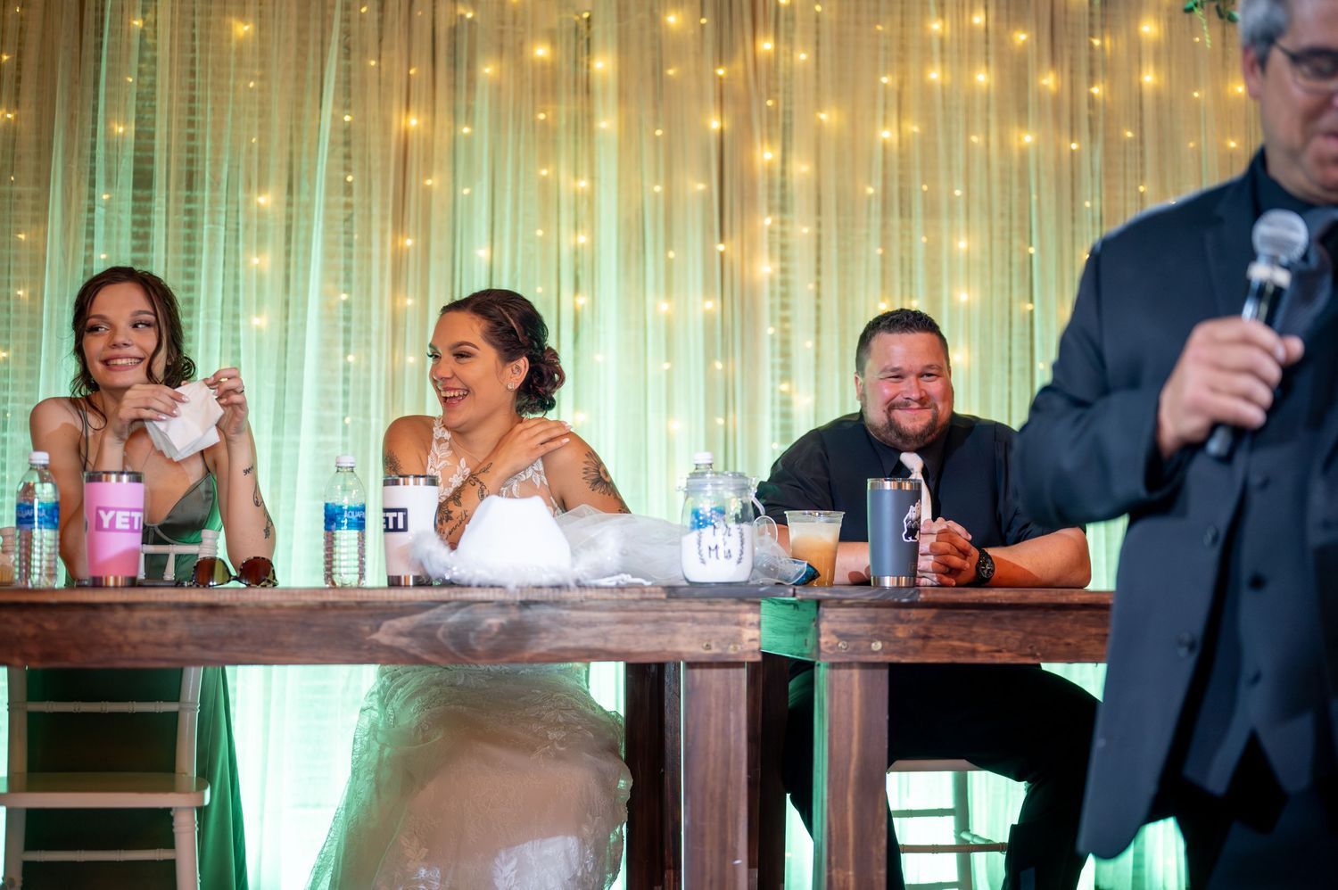 Wedding party seated at head table with string lights and mint green backdrop.