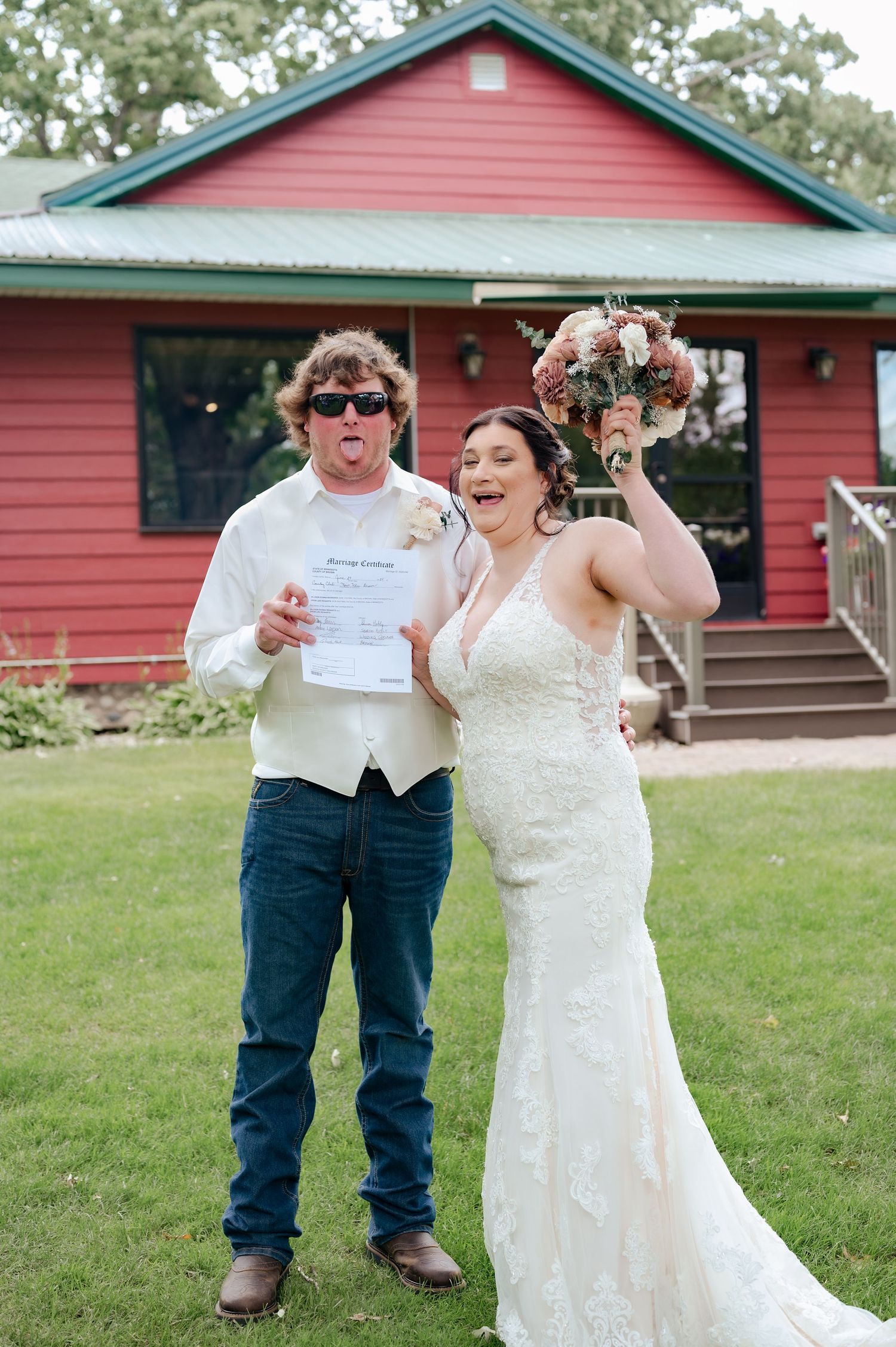 A joyful celebration outside a red farmhouse as a couple shows off their marriage certificate and bouquet.