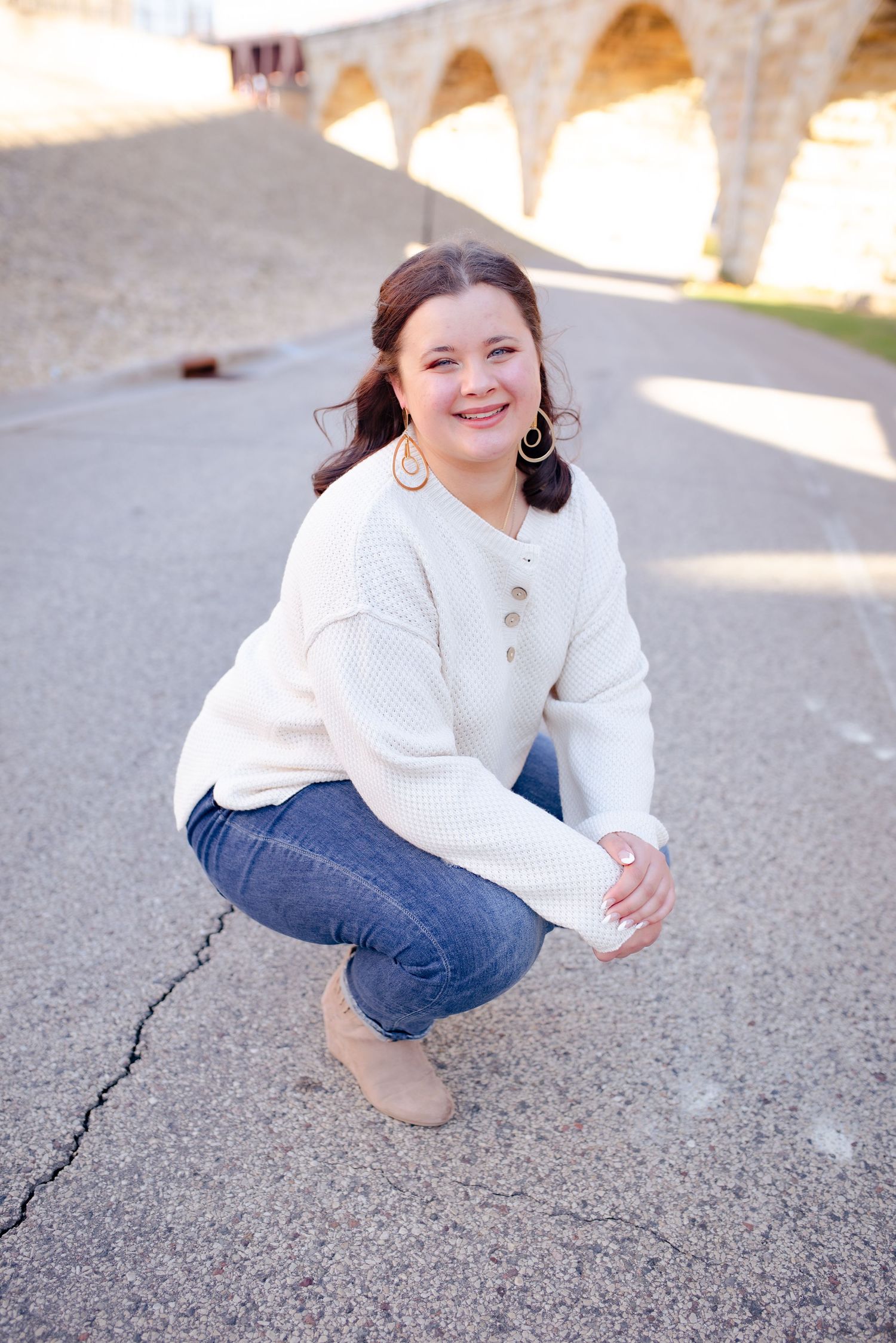 Person in white sweater and jeans crouches on pavement under stone archway.