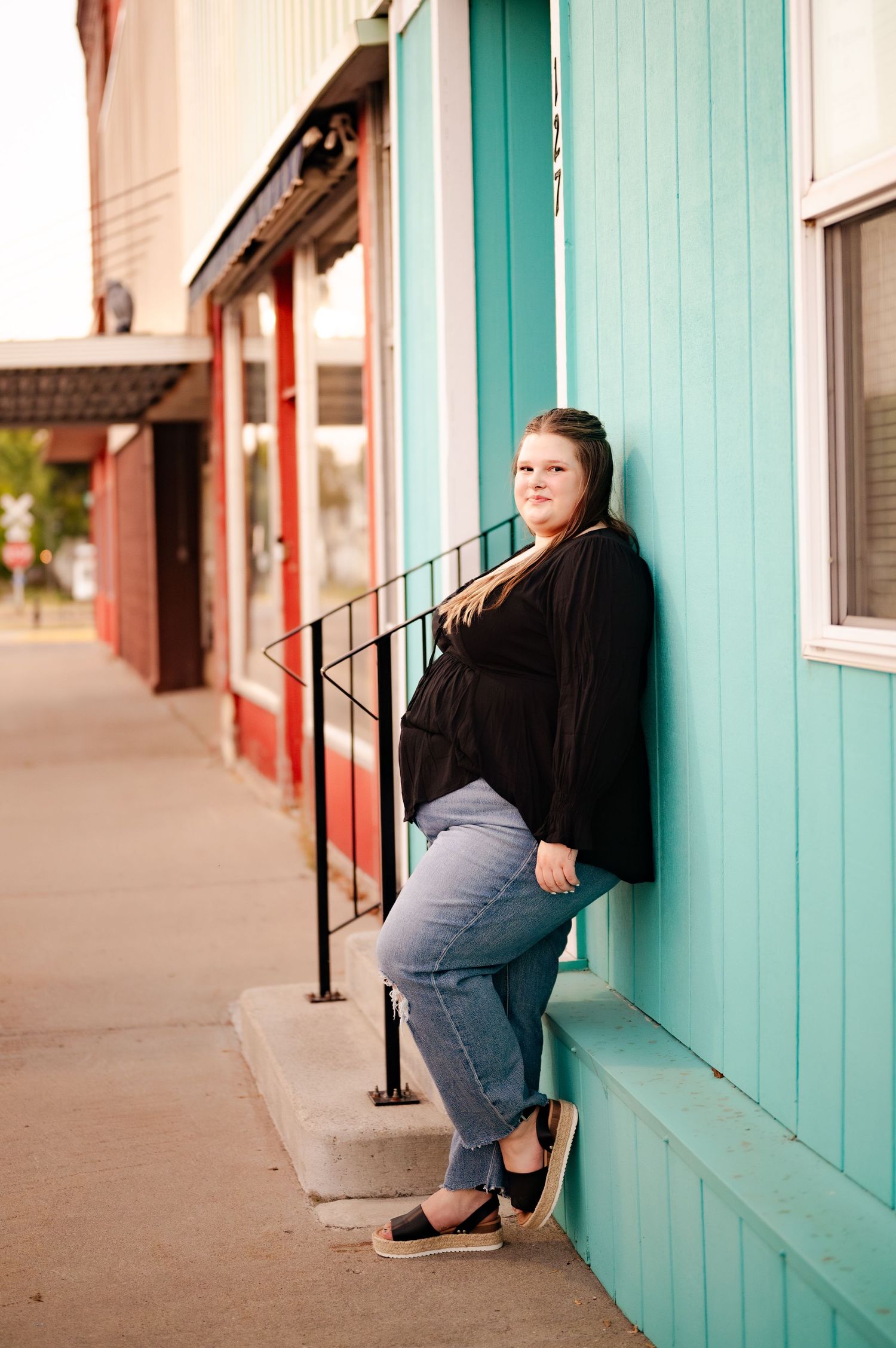 Person in black top and jeans leaning against turquoise building wall on sidewalk.