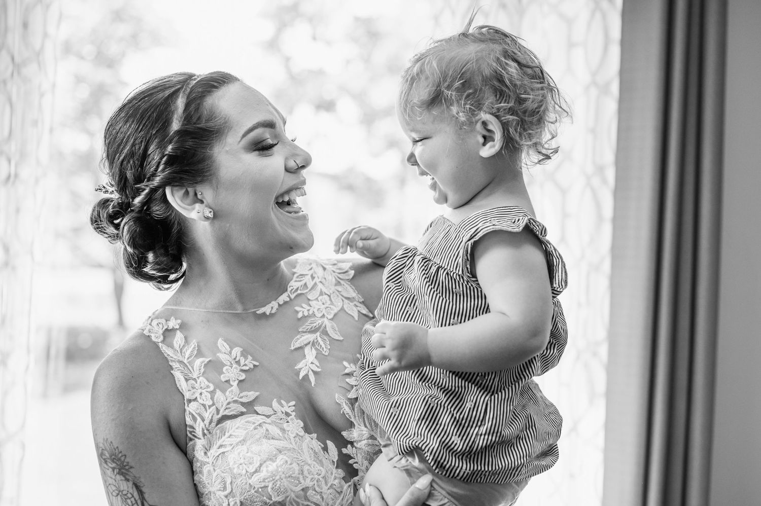 A tender moment between a bride in lace dress holding a small child against a bright window background.