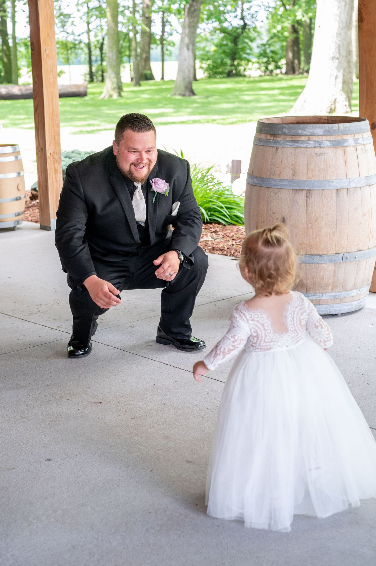 Sweet moment between groom and flower girl by rustic wine barrels at outdoor venue.