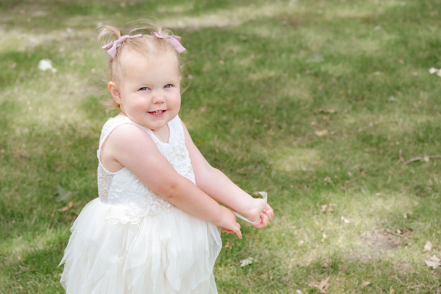 Young flower girl in white dress twirling playfully on grass during outdoor celebration.