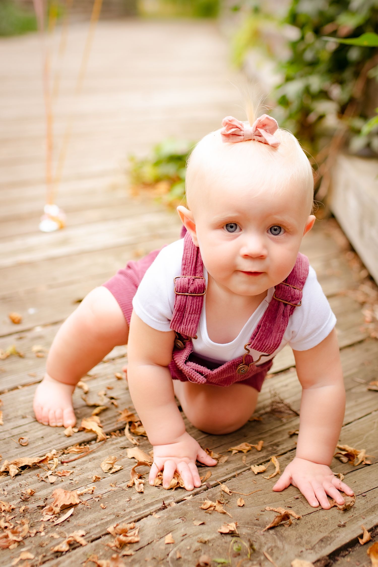 Baby in burgundy overalls crawls on wooden deck wearing pink hair bow.