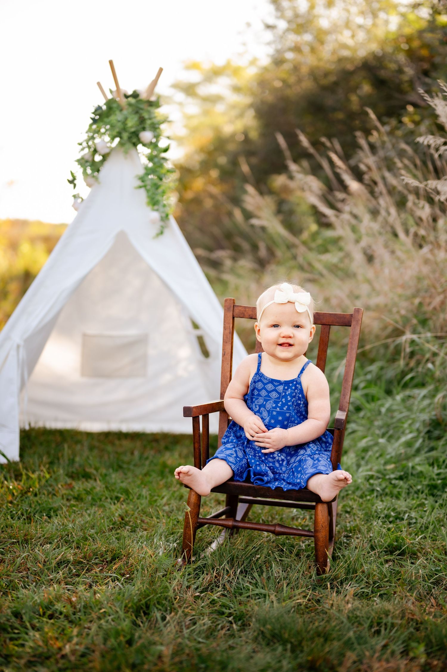 A child in a navy dress sits in a rocking chair near a decorated white teepee tent.