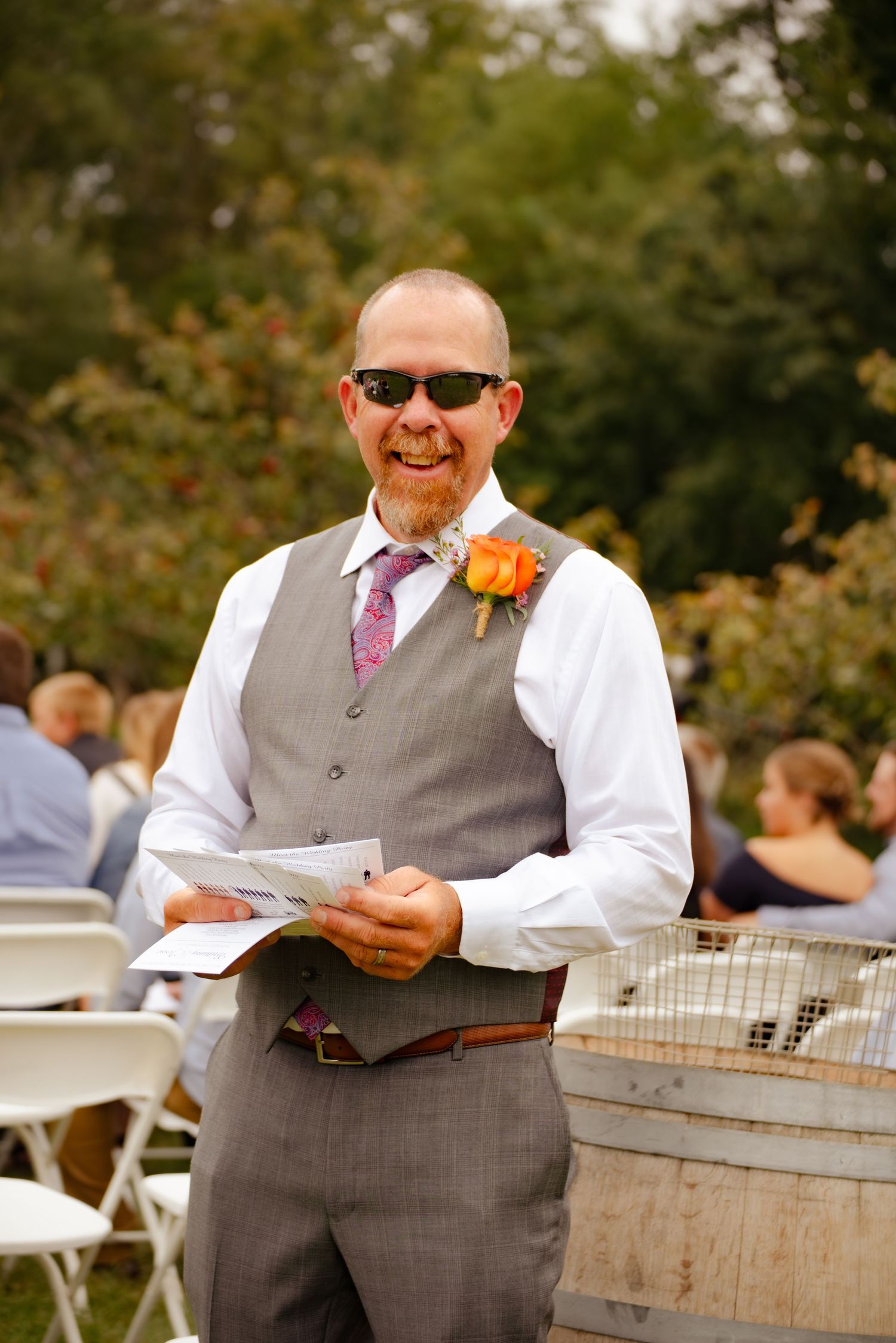 A wedding guest in a gray vest and sunglasses holds papers at an outdoor reception.