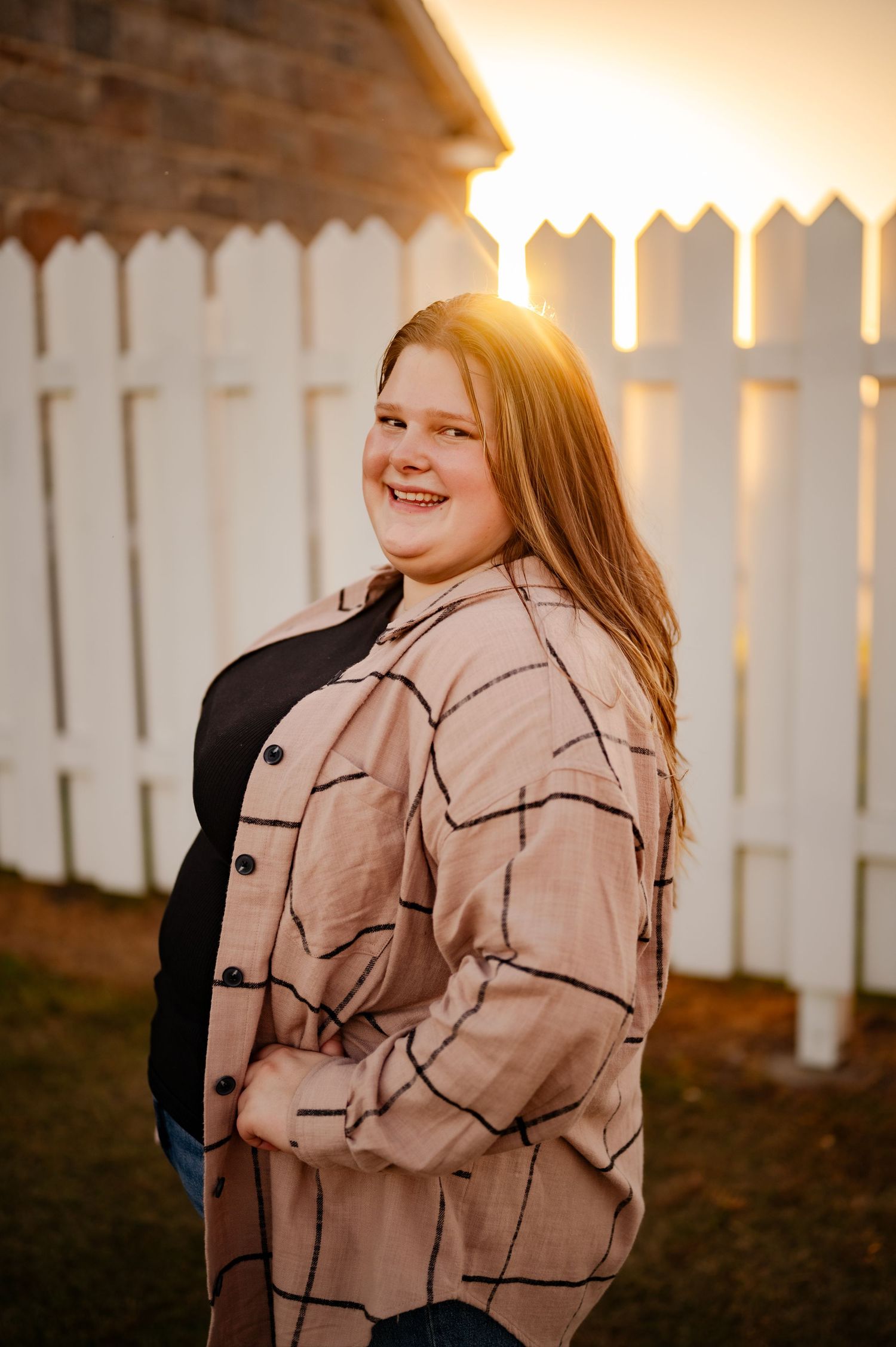 Person wearing a beige patterned jacket smiling in front of a white picket fence at sunset.