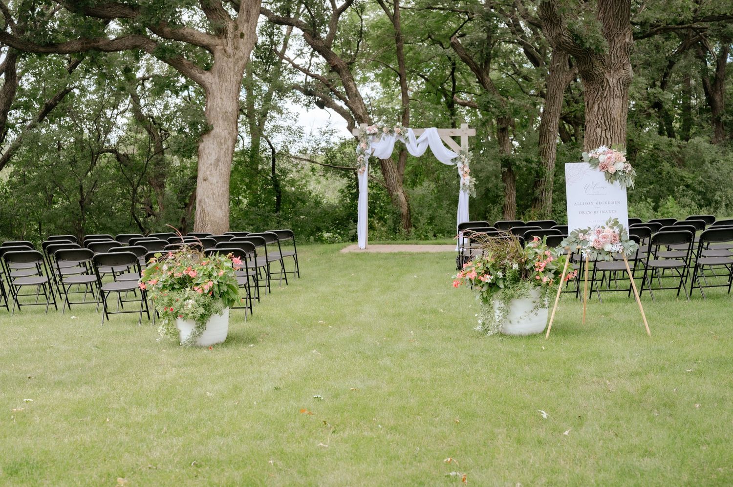 Outdoor wedding ceremony setup with white arch and folding chairs under trees.