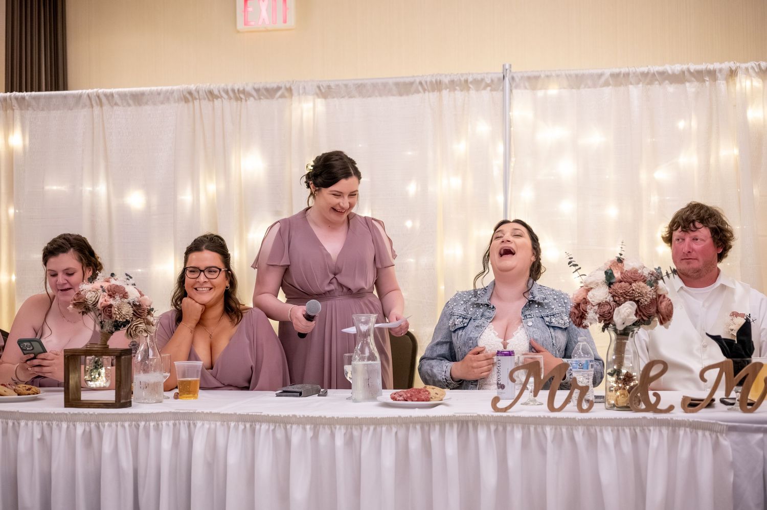 Wedding party seated at a head table with string lights and white decor in the background.