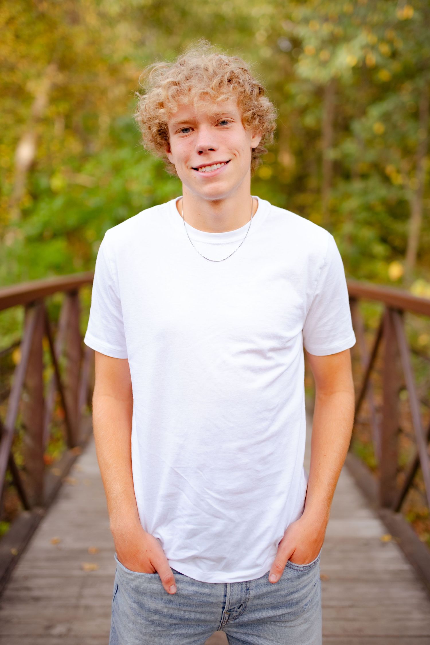 A casual portrait of someone in a white t-shirt standing on a wooden bridge surrounded by trees.