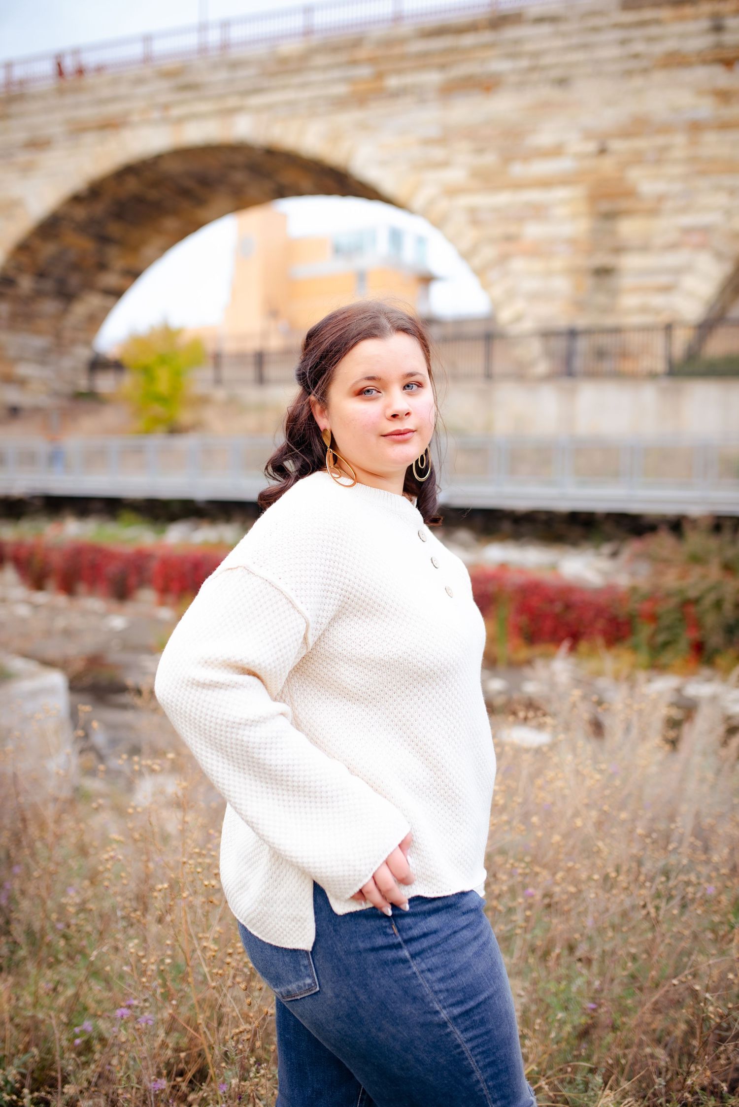 Portrait under a stone bridge with fall foliage and urban architecture in the background.