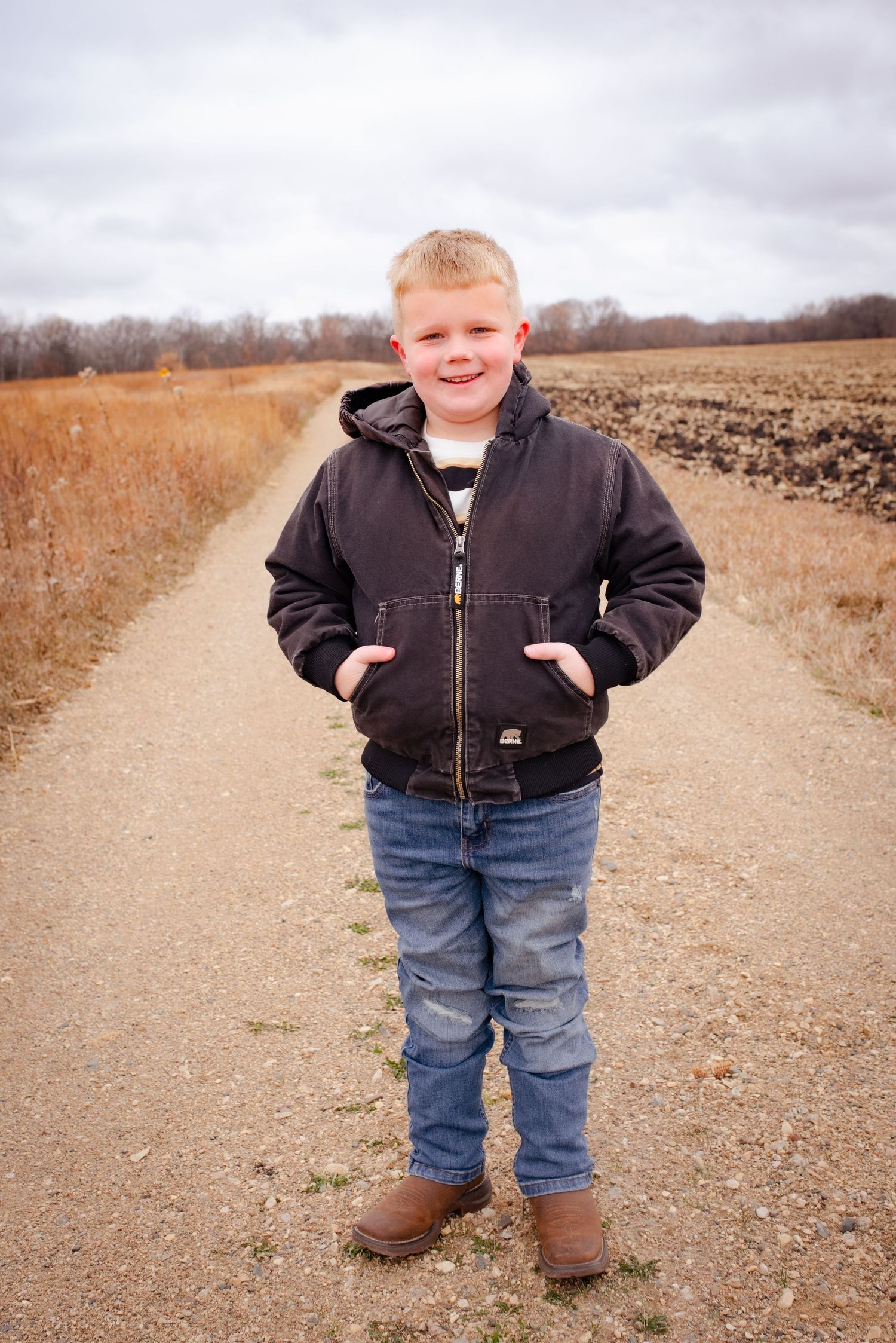 Young child stands on dirt path wearing brown jacket and jeans in rural setting.