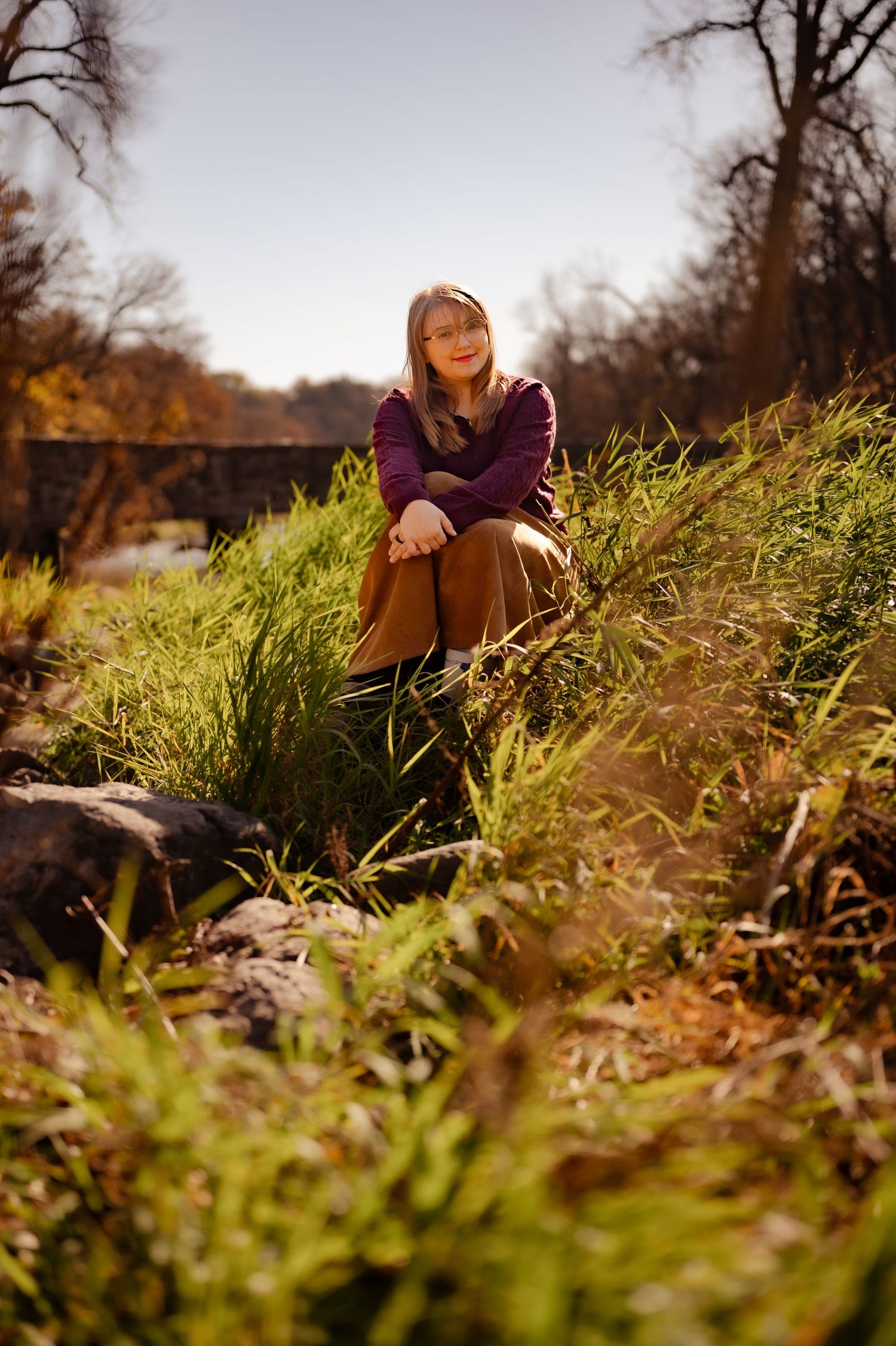 Person sits on grassy hillside in autumn wearing purple sweater and brown skirt with sunlit trees in background.