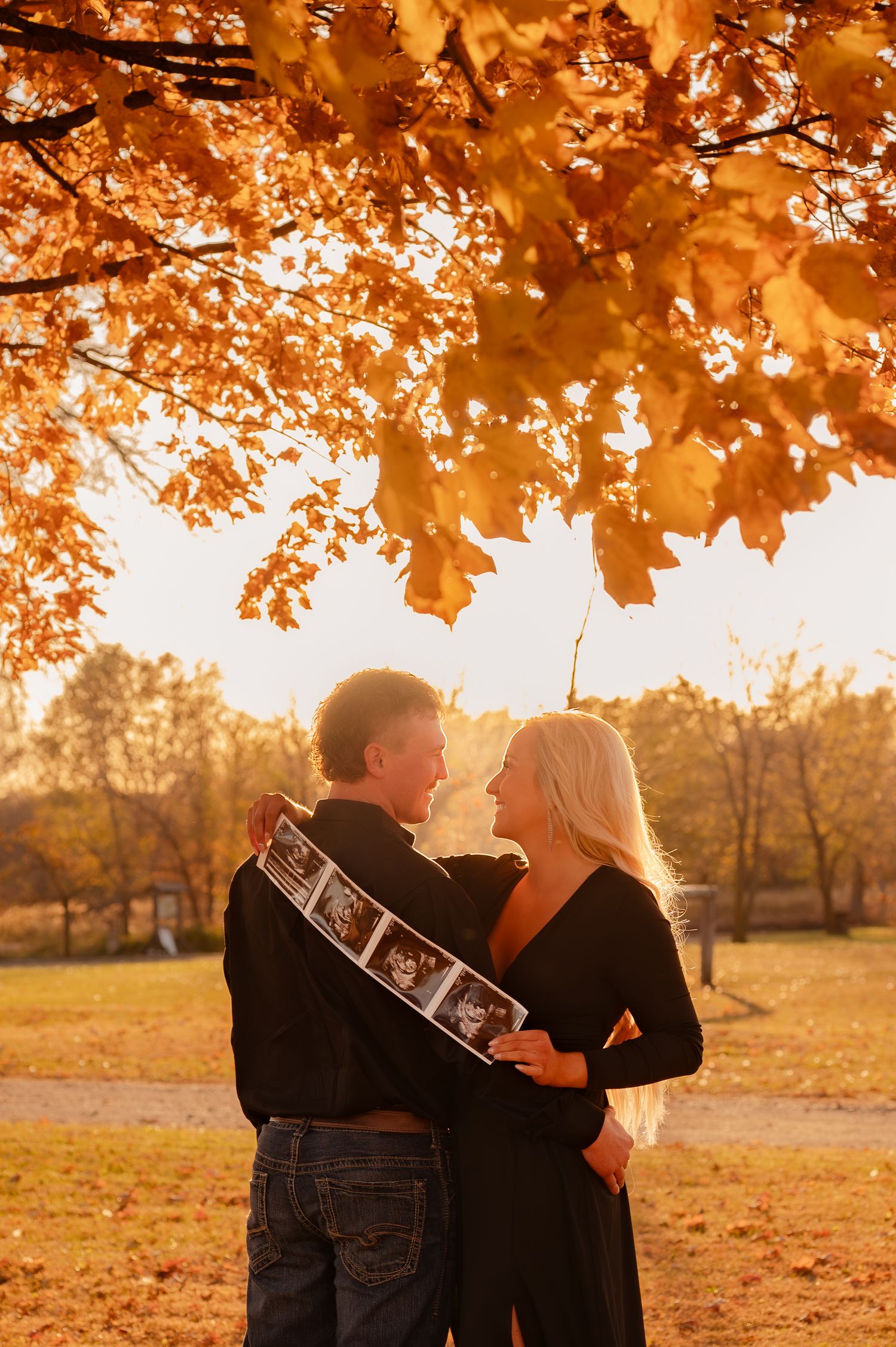 A couple shares a tender moment under golden autumn leaves while holding ultrasound photos during a fall pregnancy announcement.