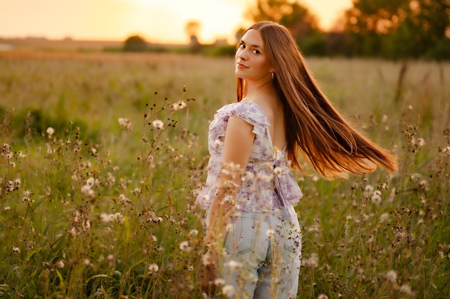 Woman in floral dress stands in golden wildflower field at sunset with long hair flowing in the breeze.