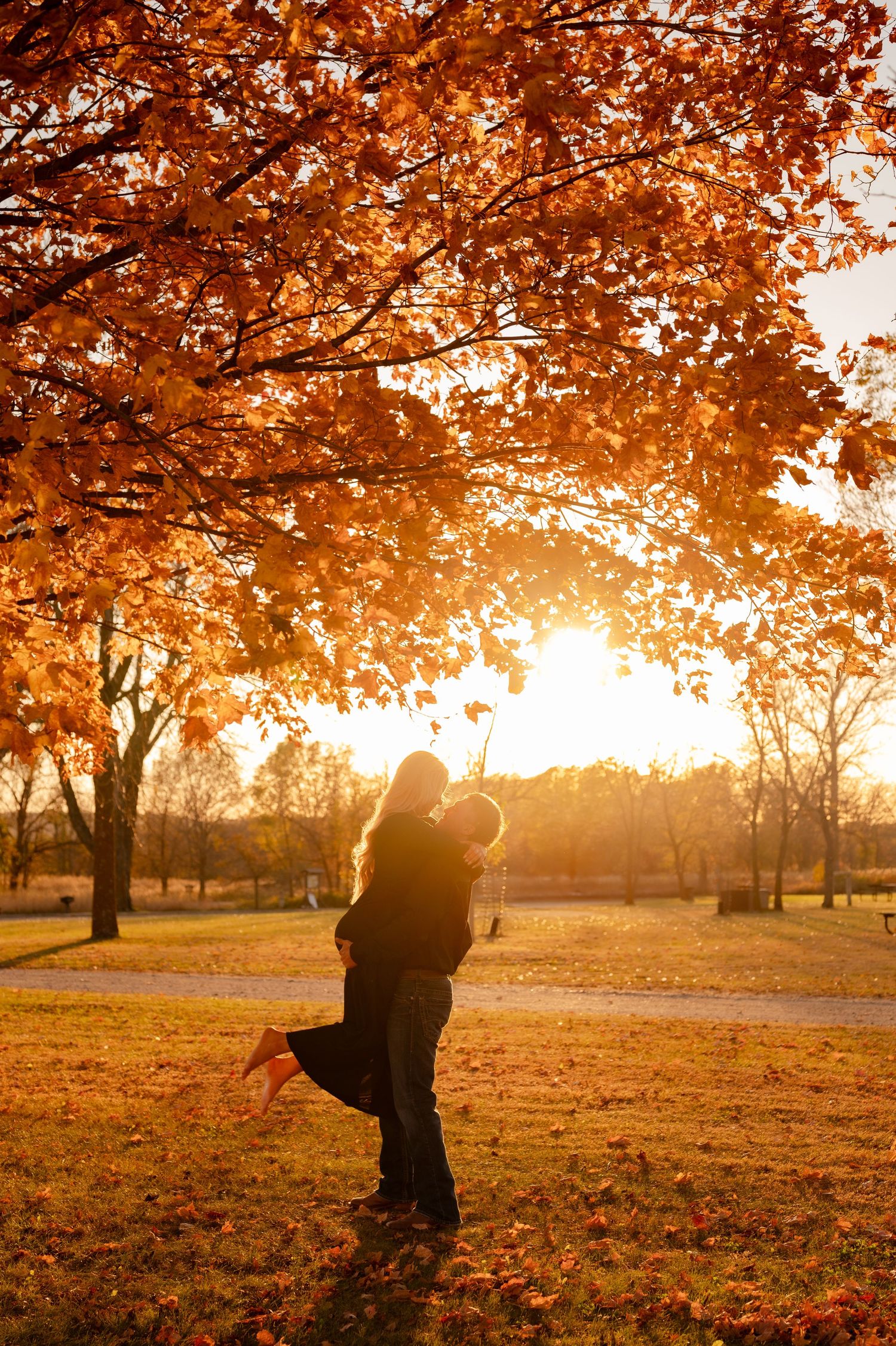Couple's silhouette against warm autumn sunset with orange leaves overhead.
