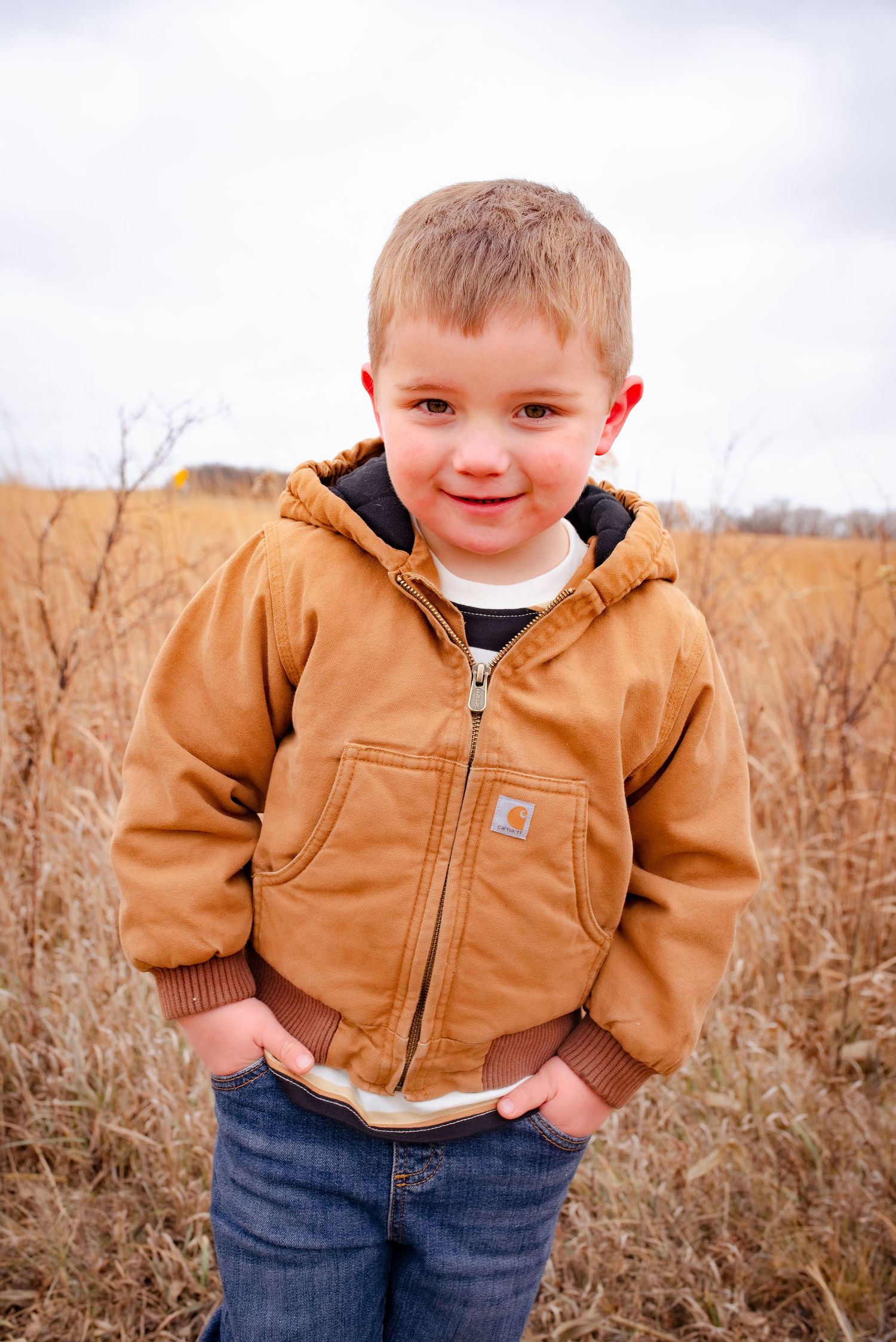 Child in brown Carhartt jacket poses in rustic field setting.