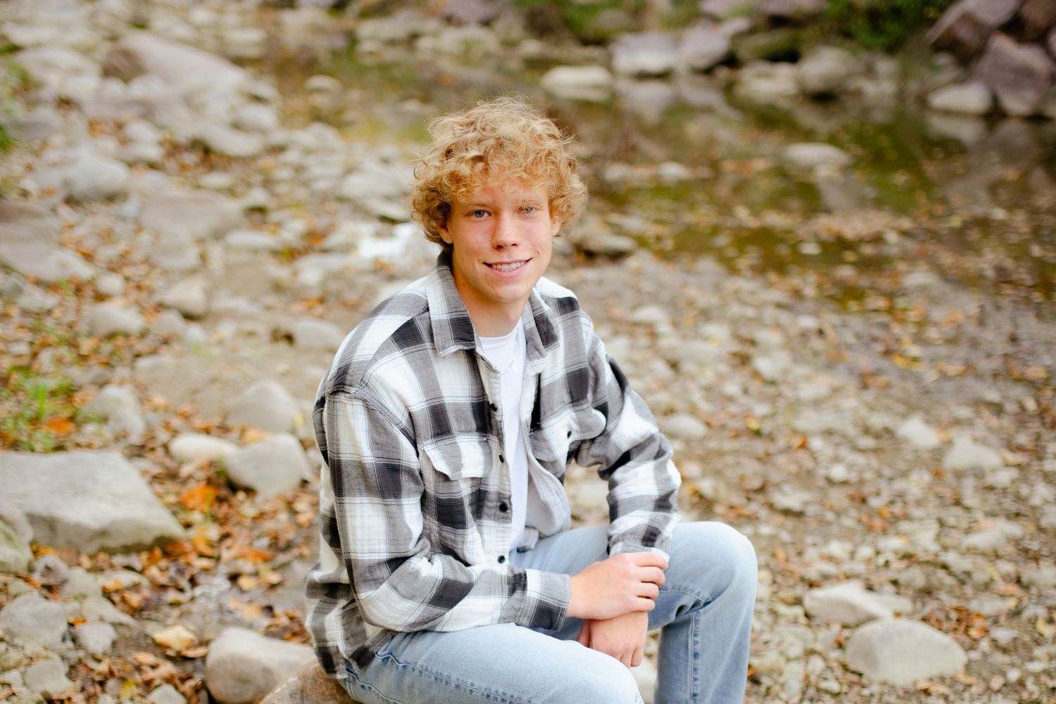 A casual outdoor portrait of someone sitting on rocks wearing a plaid flannel shirt and jeans.