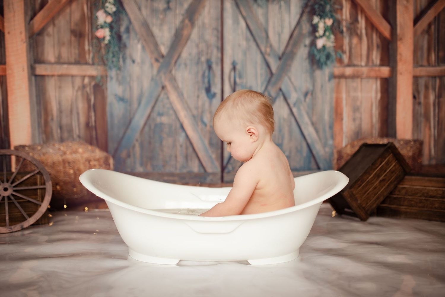 Baby sitting in vintage white bathtub with rustic barn setting and wooden crates.