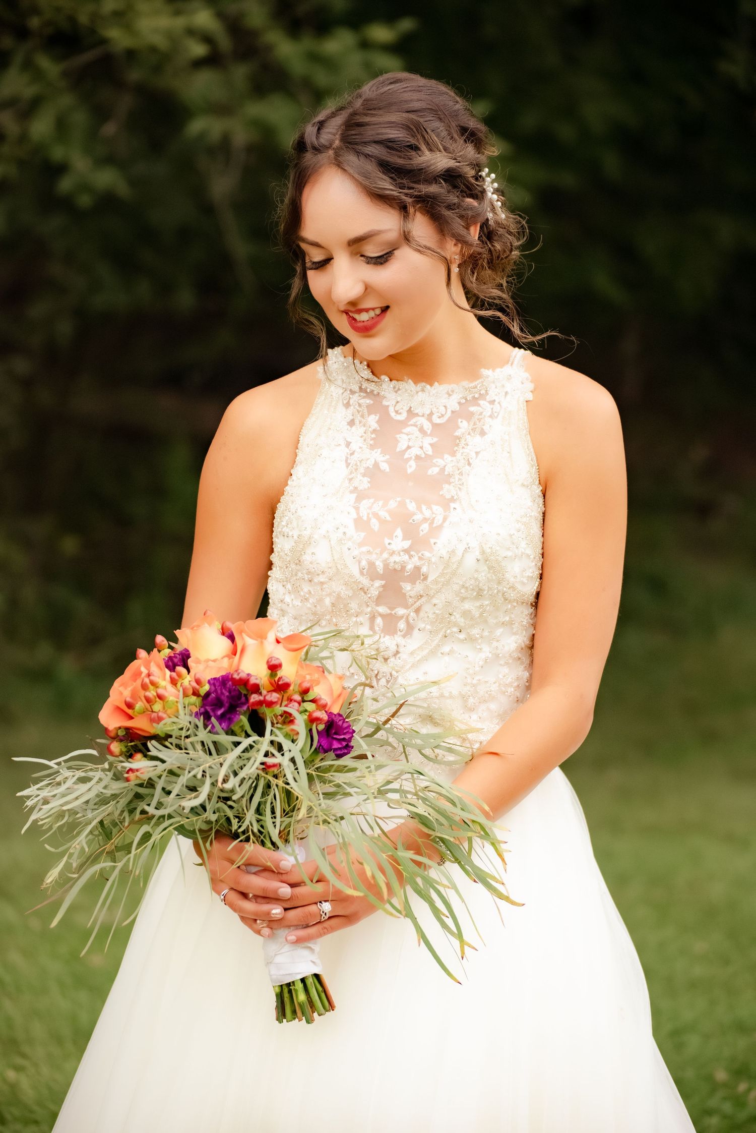 Bride in white lace dress smiles while holding colorful bouquet of orange and purple flowers.