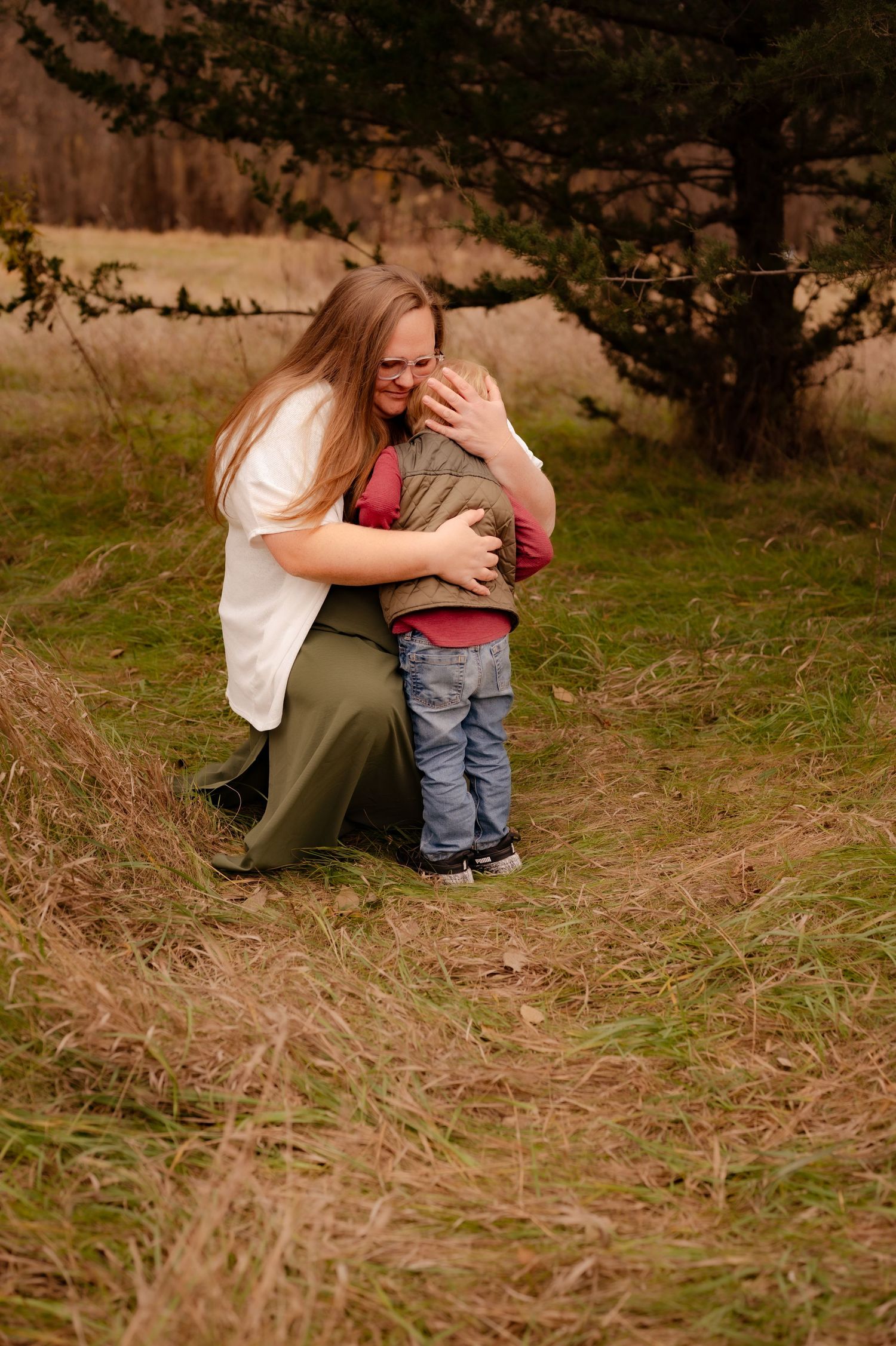 A tender embrace shared between two people in a grassy field during golden hour with a tree in the background.
