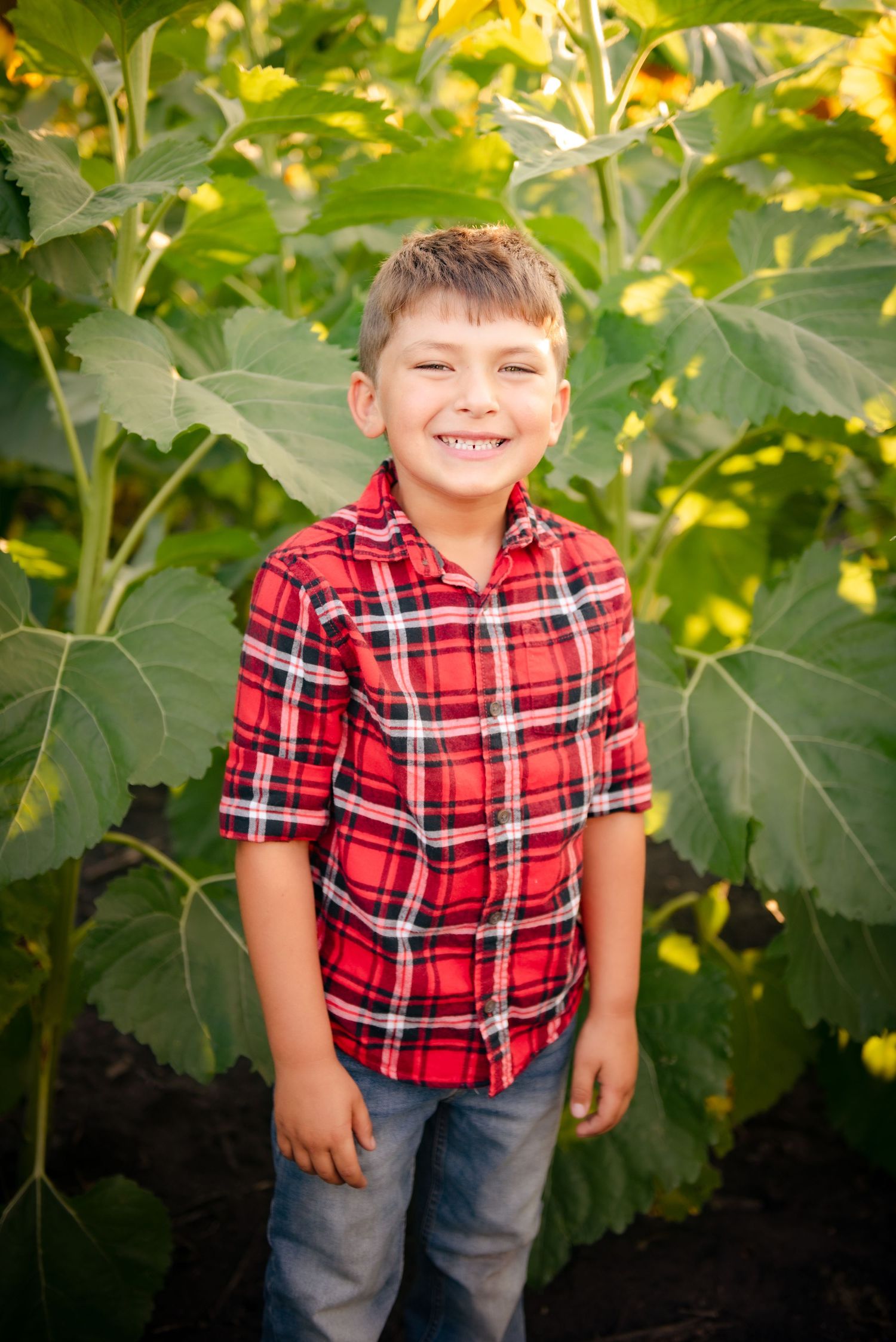 Young person in red plaid shirt smiling brightly against backdrop of large green leaves.