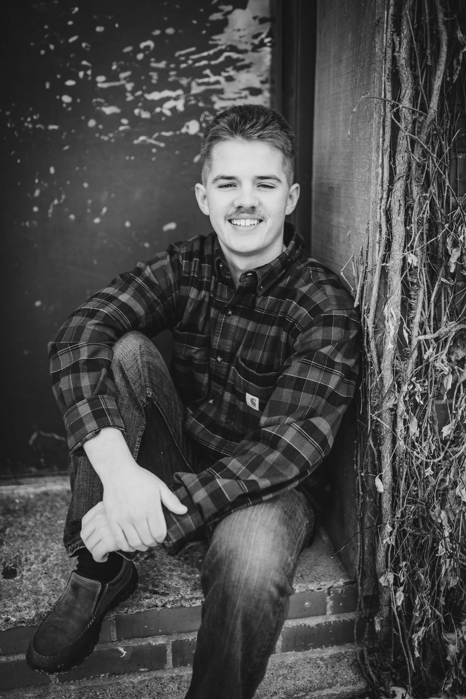 Black and white portrait sitting by vintage door wearing plaid flannel shirt.