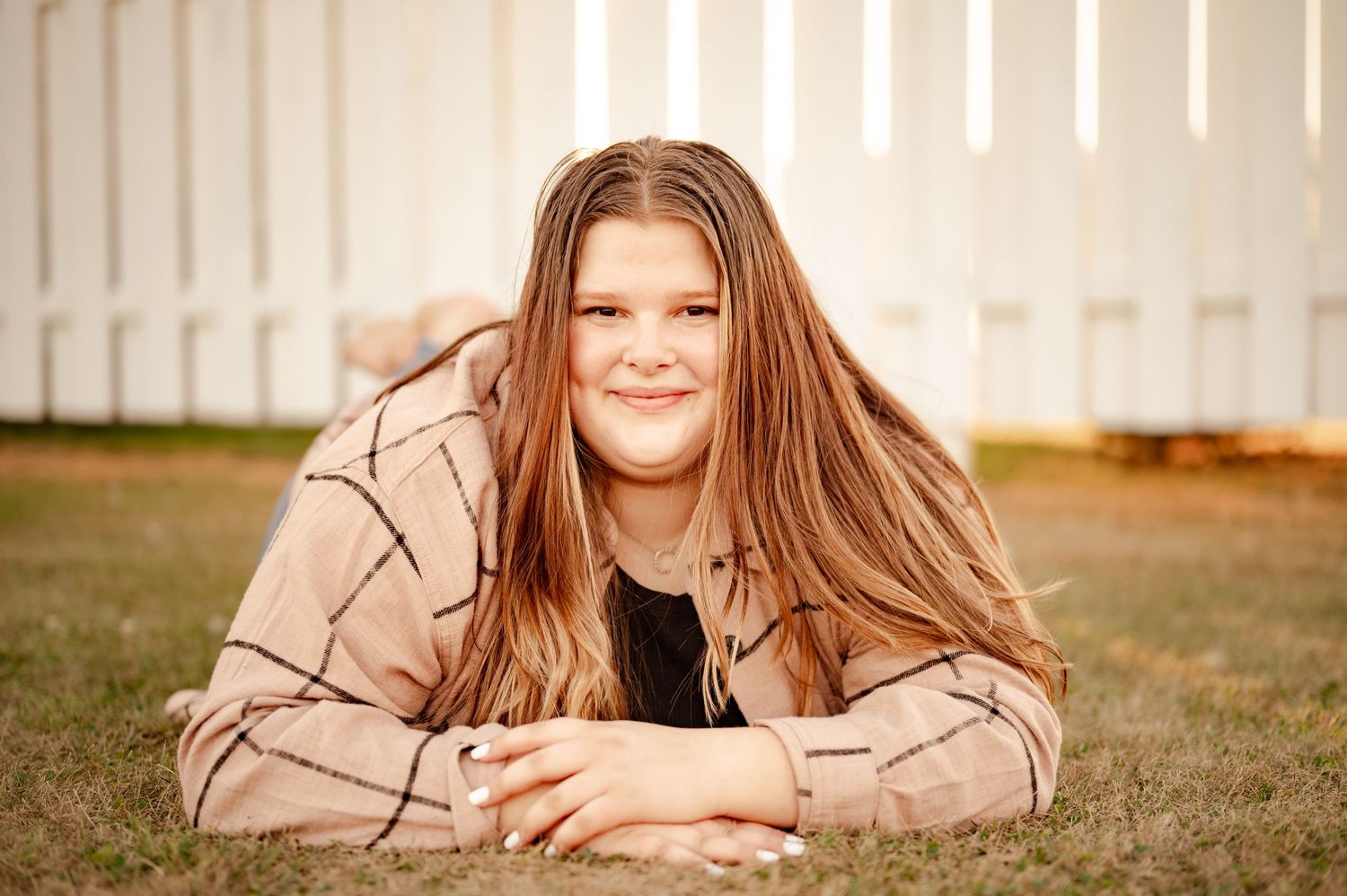 A casual outdoor portrait on grass with a plaid jacket against a white fence background.