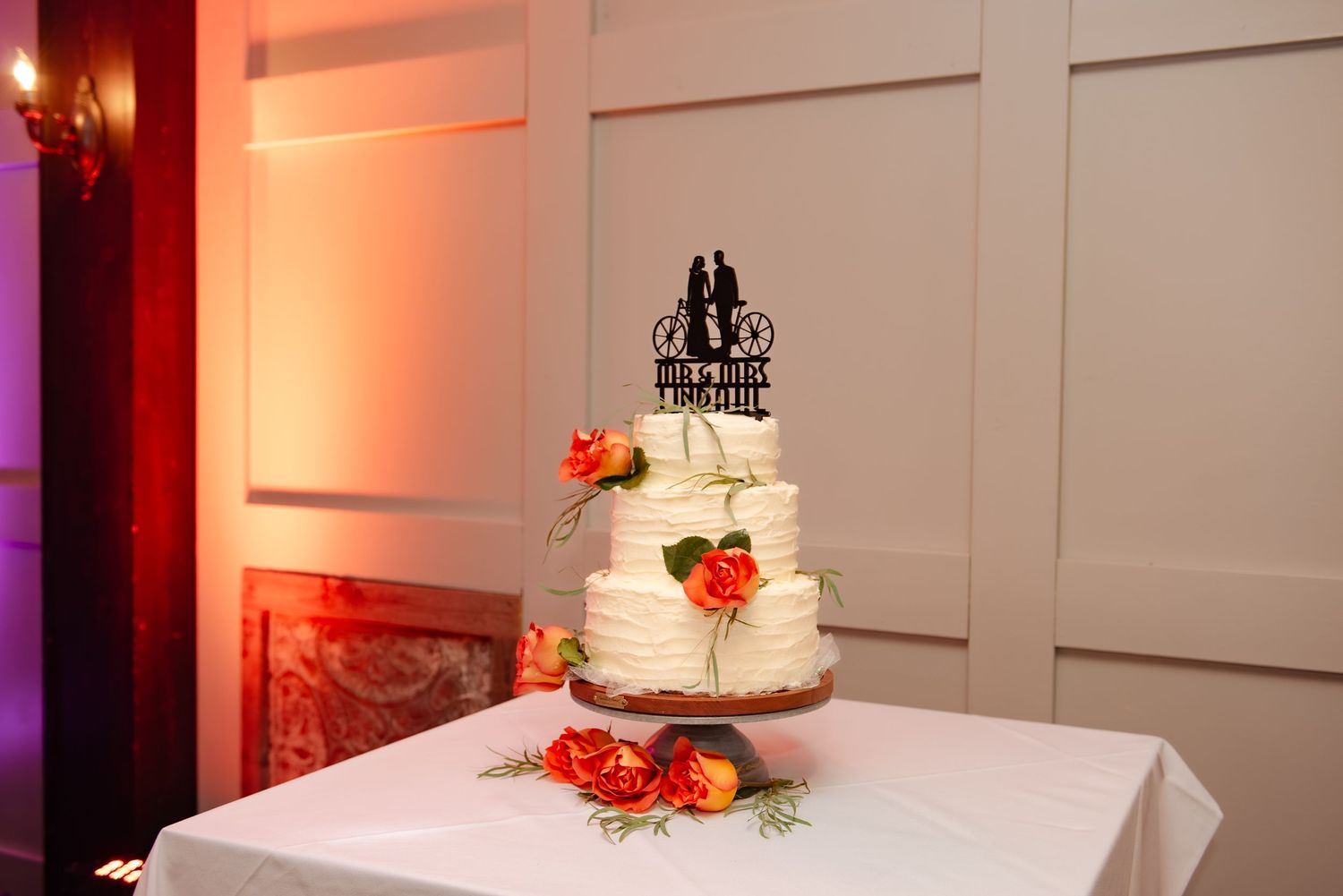 White wedding cake with red flower decorations displayed on a table with dramatic lighting.