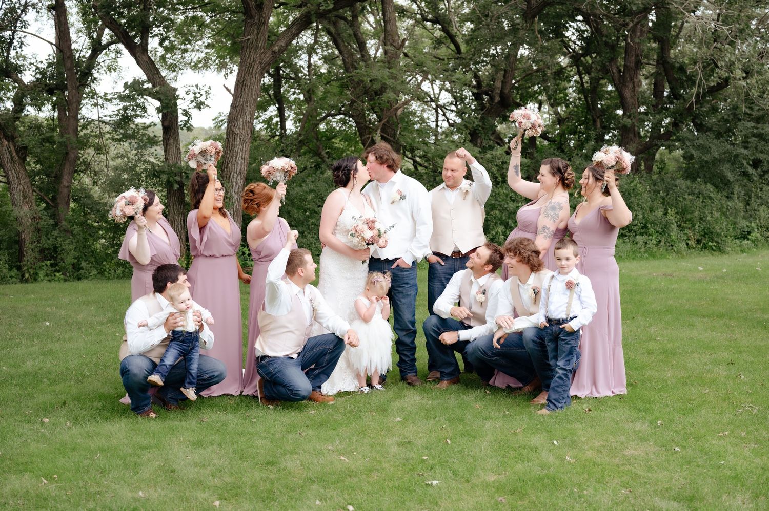 Wedding party in pink dresses and casual attire poses playfully on grass with trees in background.