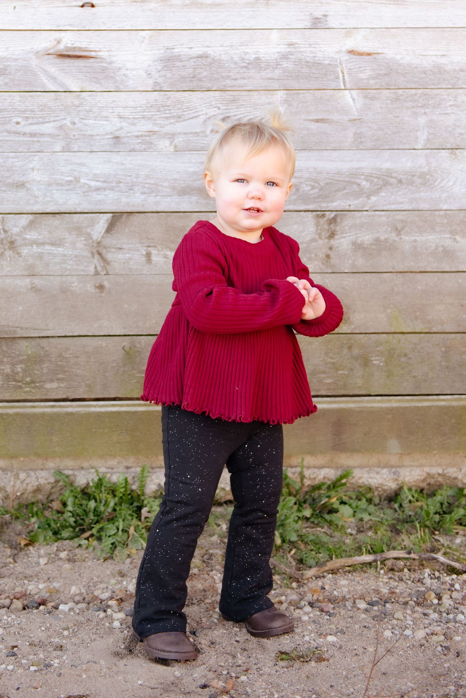 A toddler wearing a red sweater dress and dark pants stands against a rustic wooden wall.