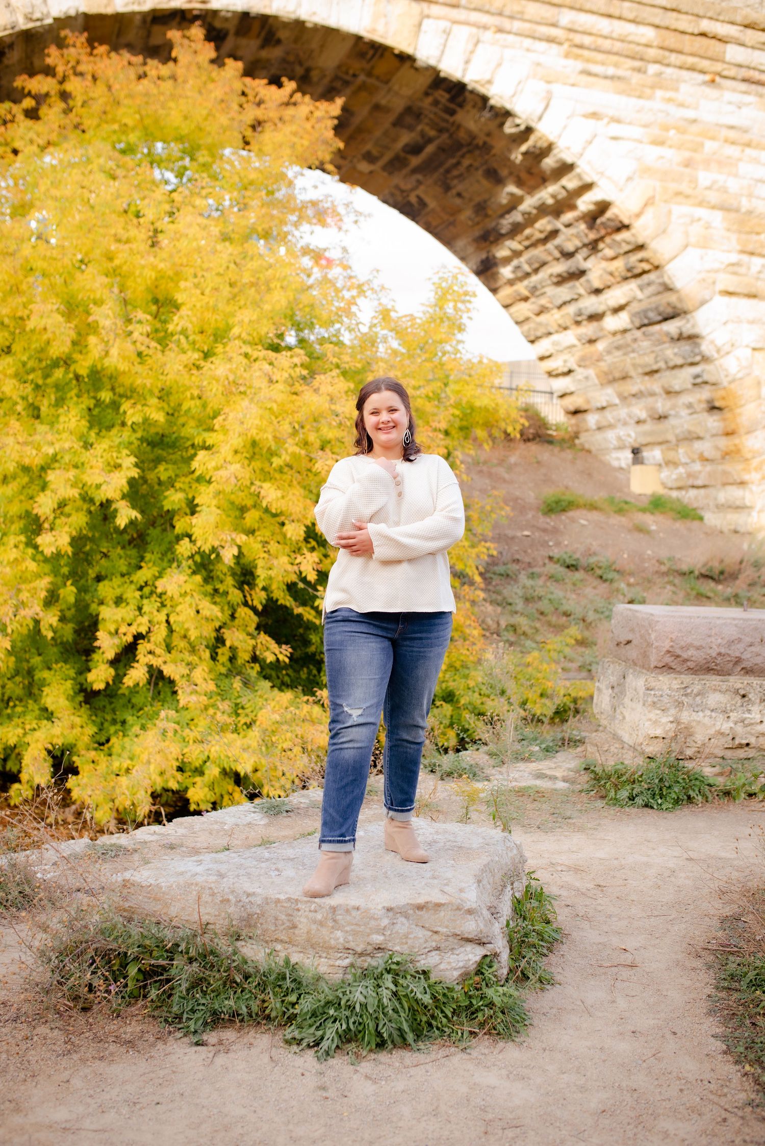 Person in cream sweater stands on stone platform under bridge arch with yellow autumn foliage.