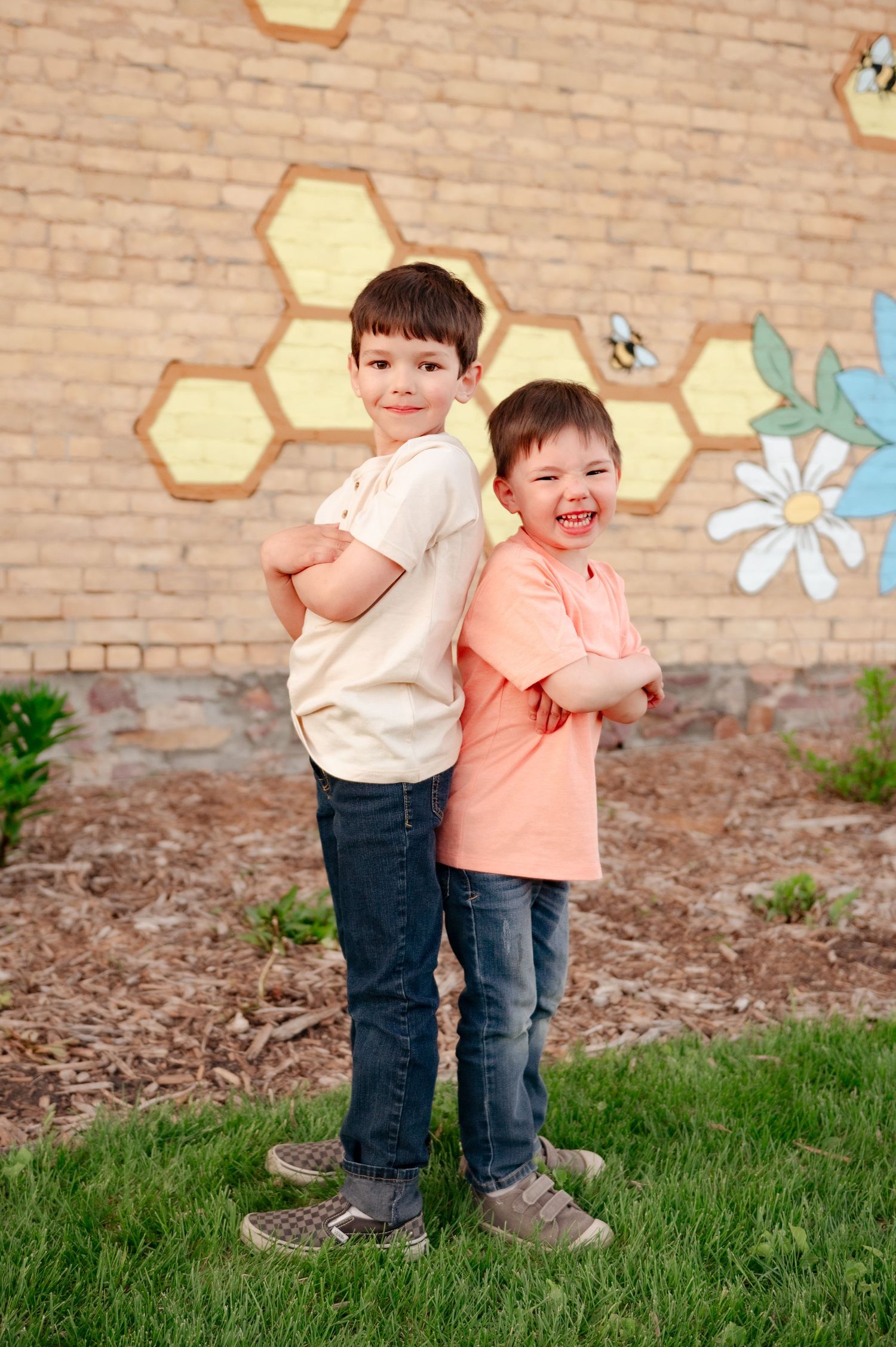 Two children pose back-to-back outdoors in coordinating casual outfits with painted wall background.
