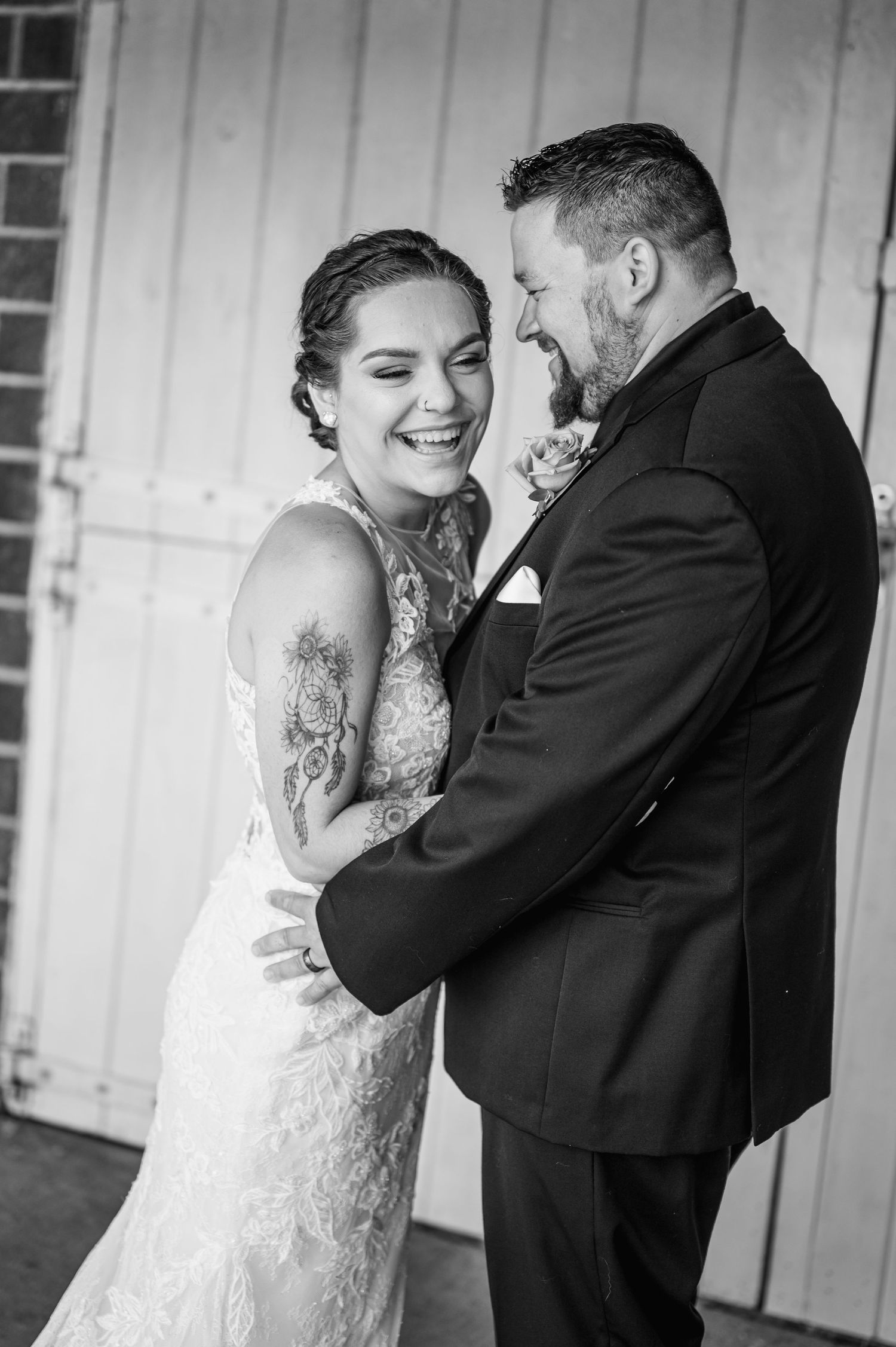 Black and white portrait of laughing newlyweds in tender embrace by rustic door.