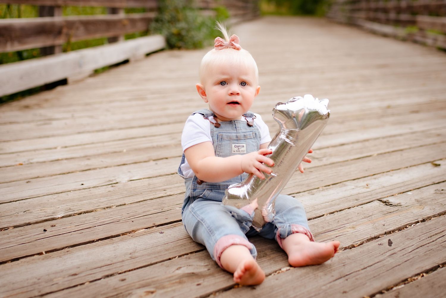 Baby wearing overalls sits on wooden boardwalk holding a silver number decoration.