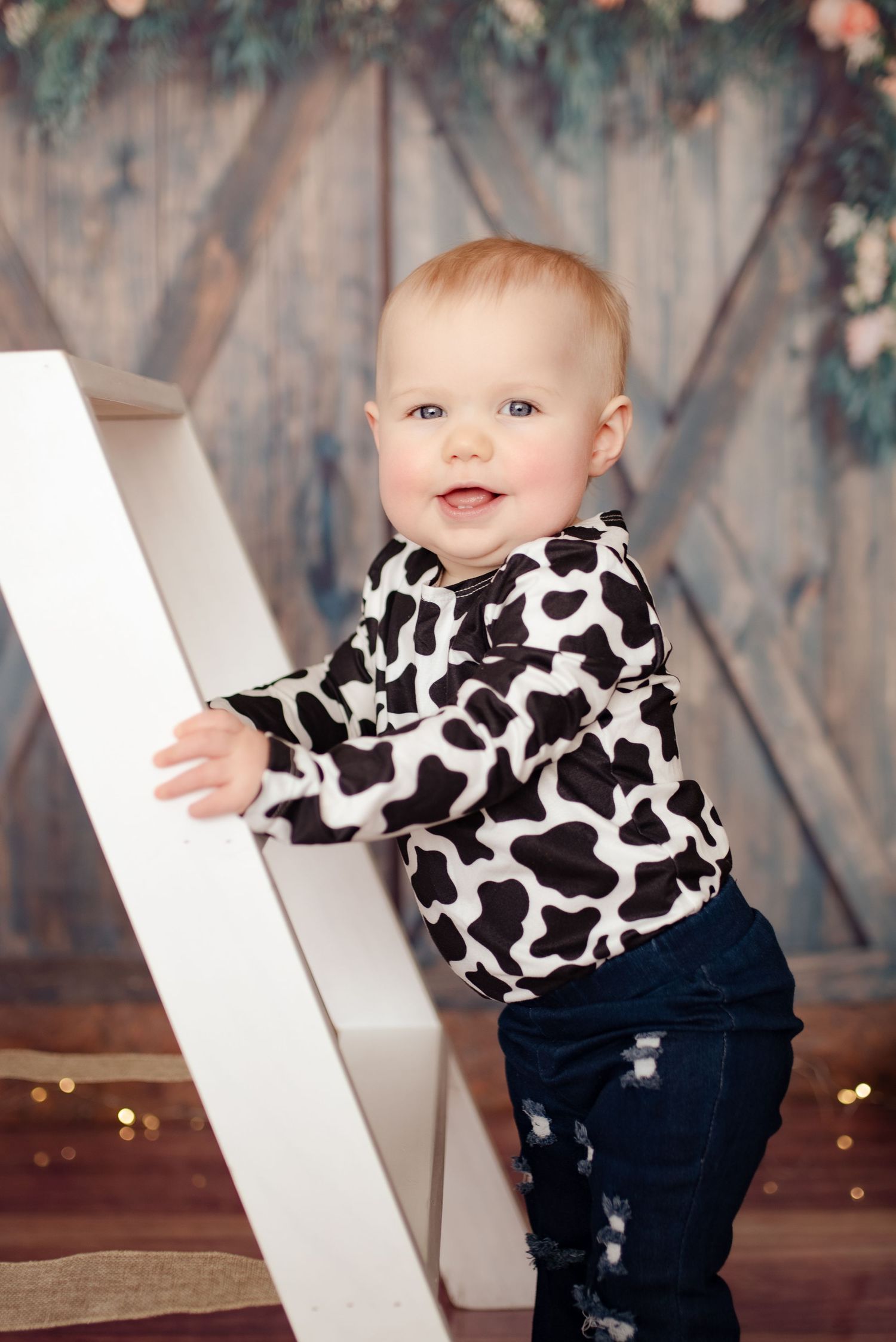 Baby in cow print sweater stands on white ladder against floral backdrop.