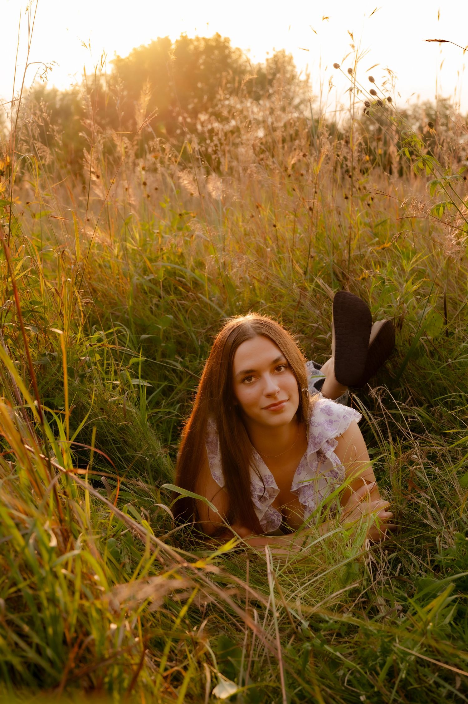 A woman sits among tall grass and wildflowers in warm evening sunlight.
