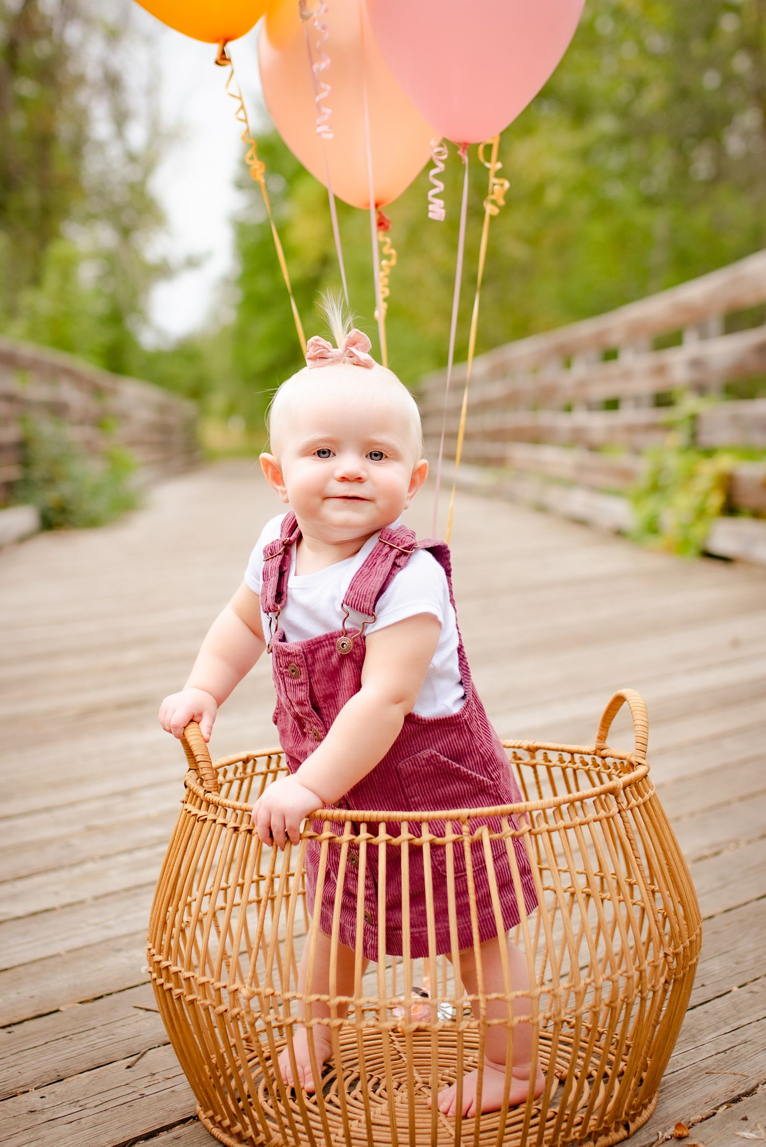 Baby in burgundy overalls sits in wicker basket with pink and orange balloons on wooden bridge.