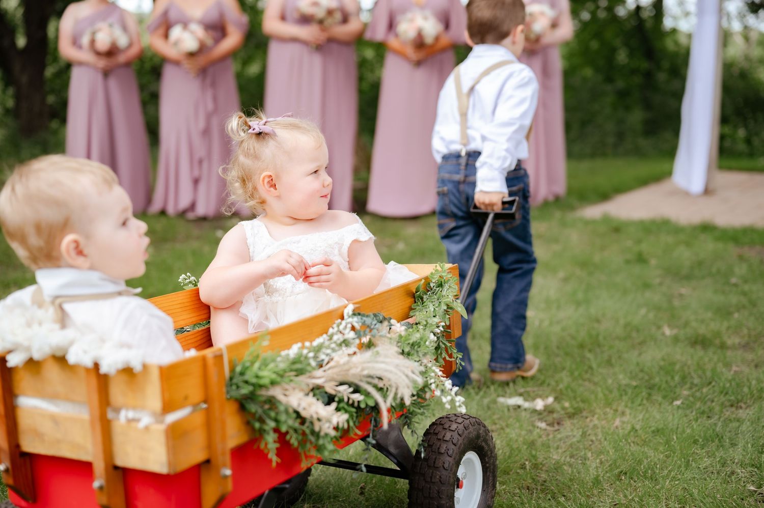 Small children seated in a wooden wagon with flowers at an outdoor celebration.