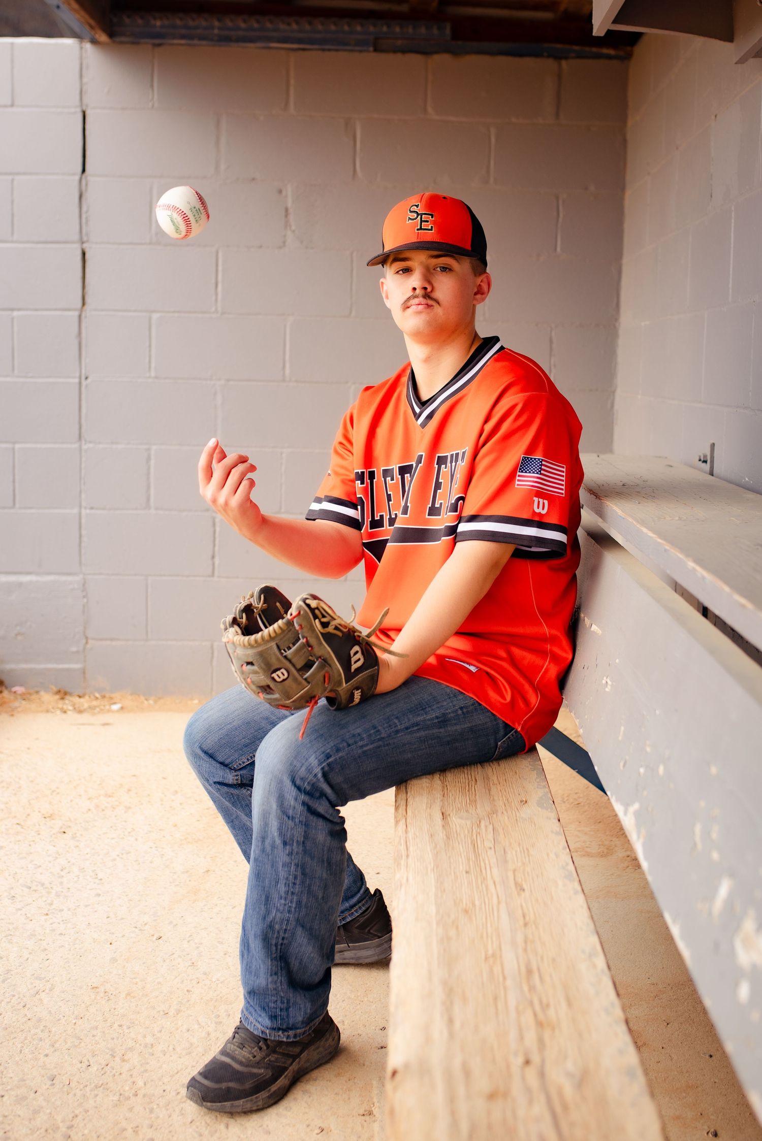 Baseball player in orange uniform tosses ball while sitting in dugout.