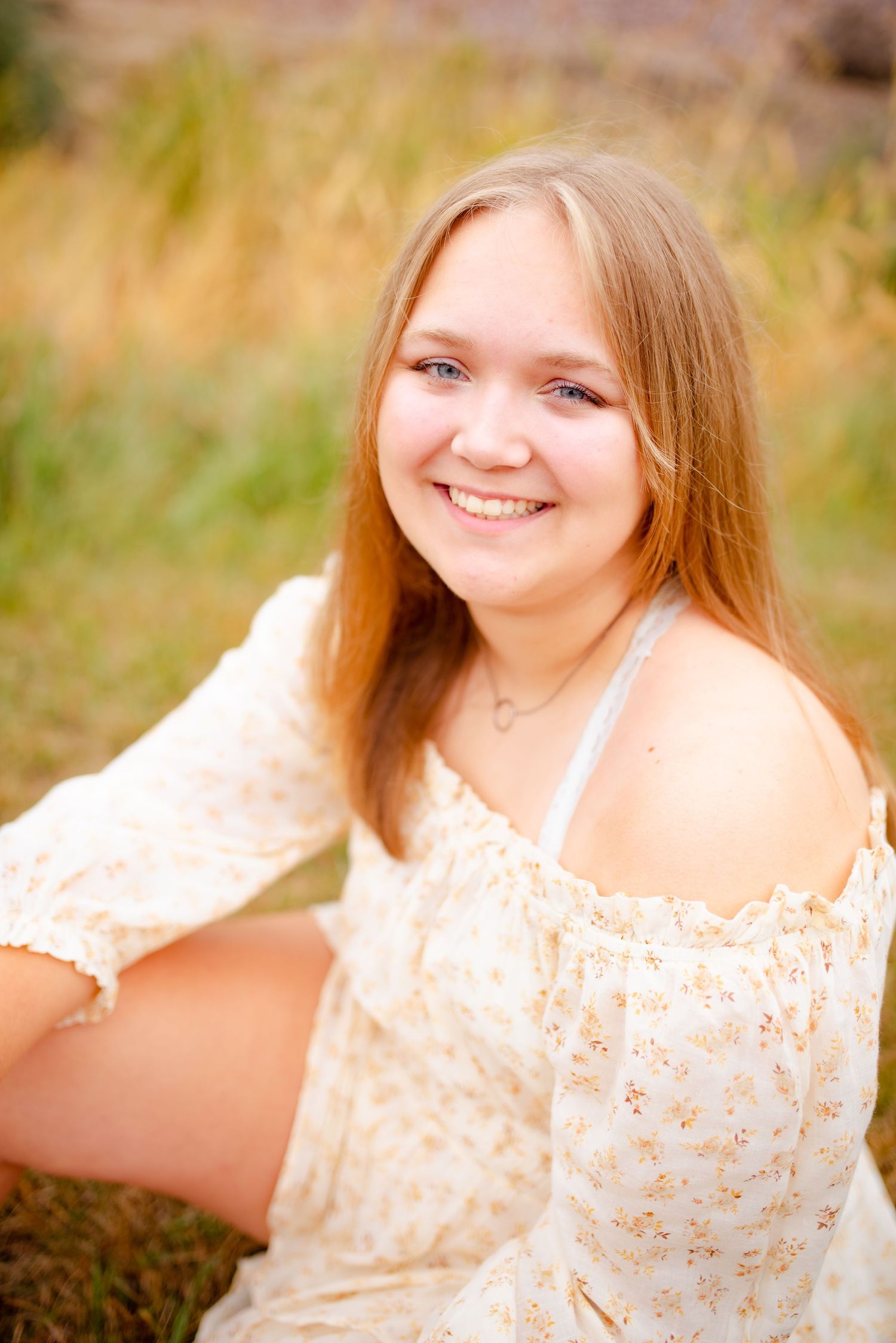 Outdoor portrait of person in white floral dress sitting in golden field at sunset.