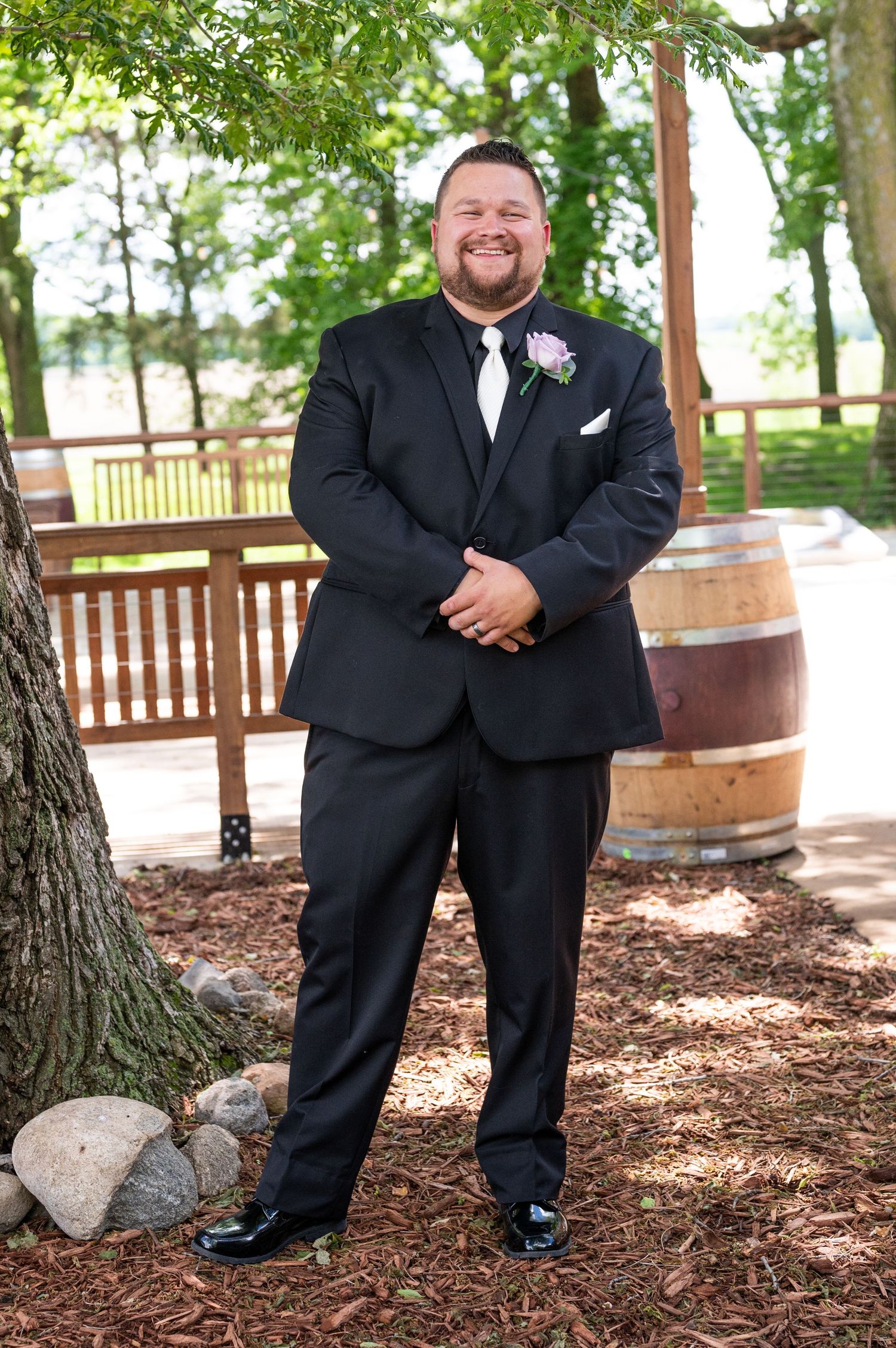 Groom in black tuxedo poses outdoors near rustic wine barrels and trees.
