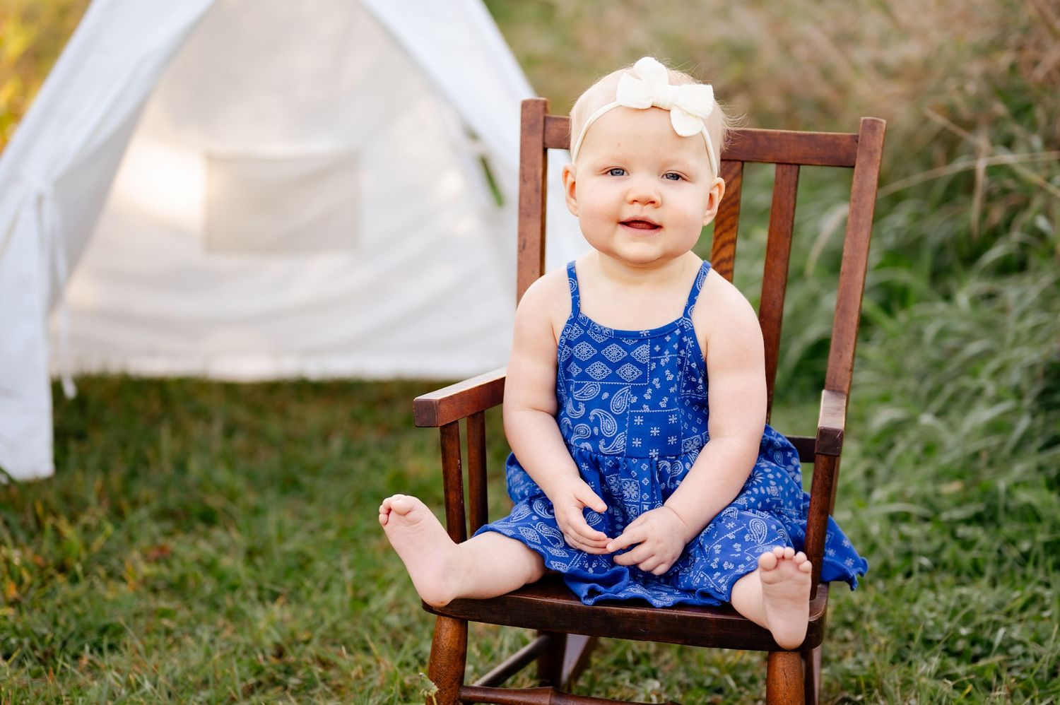 A blue sundress-clad toddler sits in a wooden rocking chair with a white tent in the background.