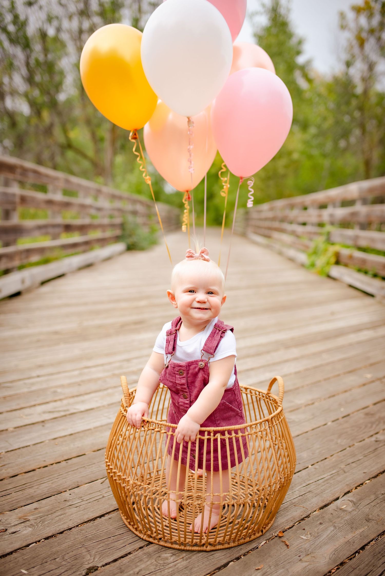 Baby in red overalls sits in wicker basket with colorful balloons on wooden boardwalk.