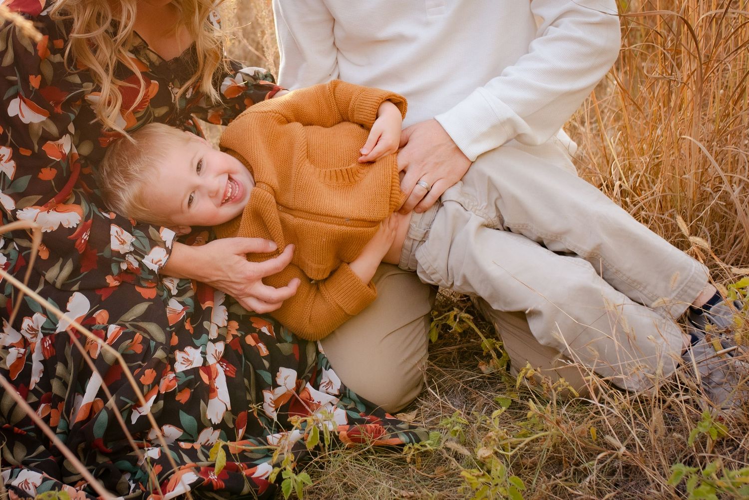 A toddler in an orange sweater lies playfully in autumn grass during an outdoor family photoshoot.