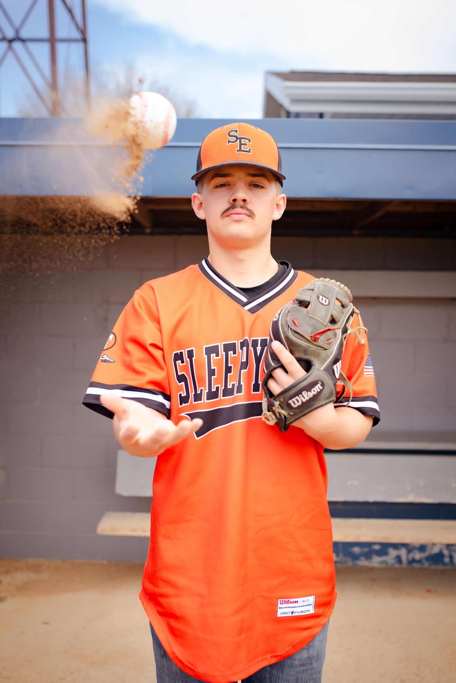 Player in orange baseball uniform holds glove as dirt and dust swirl in dramatic lighting.