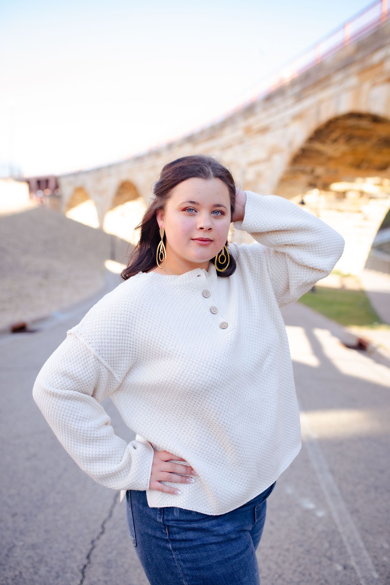 Person in white sweater posing near stone bridge arch in sunlight.