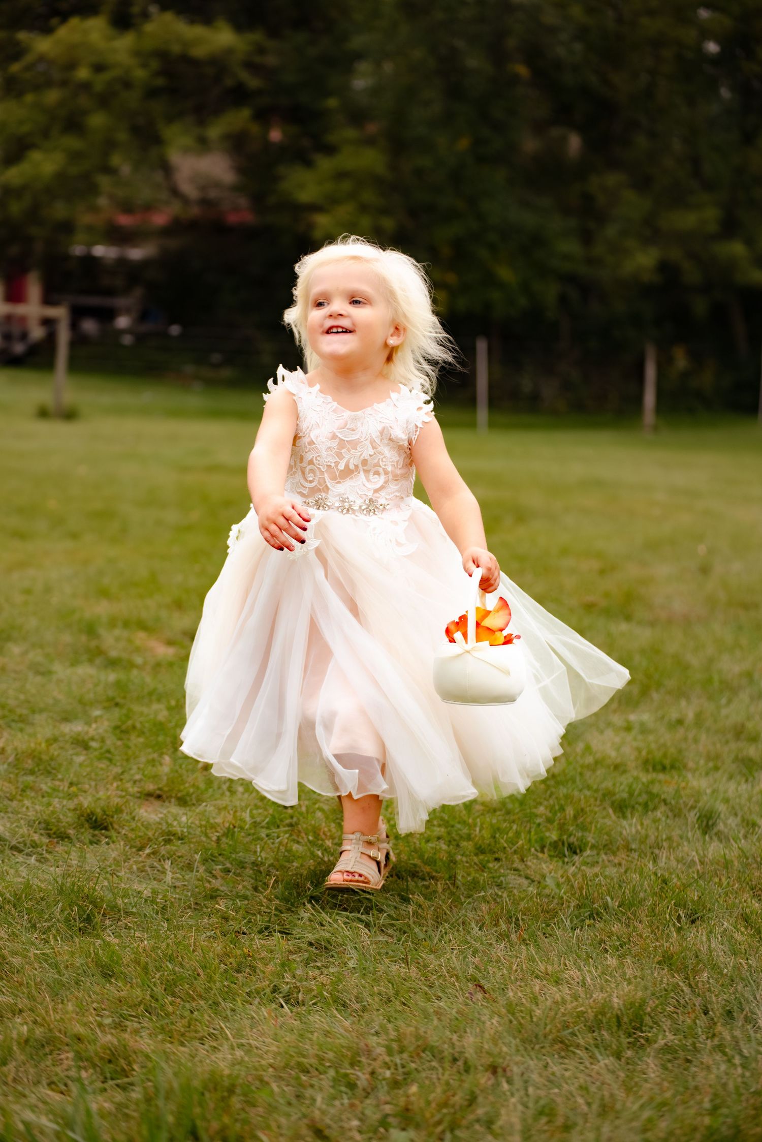 A flower girl in a lace and tulle dress dances joyfully while holding a white basket outdoors.