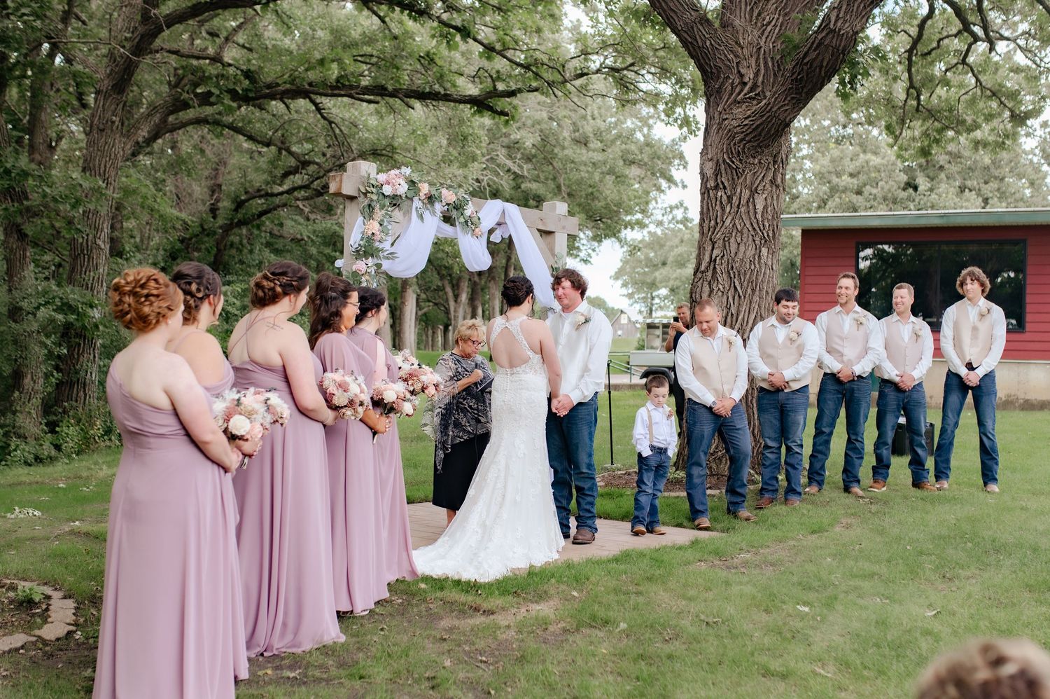 Wedding party in pink dresses and jeans lined up outdoors under trees.